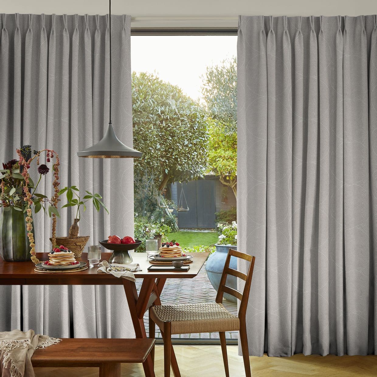 Pleated floor-to-ceiling light-gray curtains, parted to reveal a glass patio door, admitting soft daylight into a dining area with a wooden table set for breakfast and a leafy garden beyond.