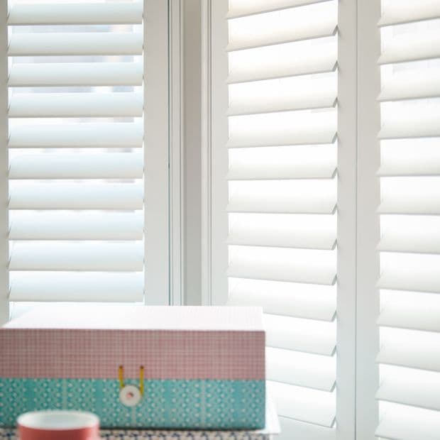 White plantation shutters with wide horizontal slats cover corner windows, closed and filtering bright daylight; low surface in foreground holds patterned storage boxes and a small cup.