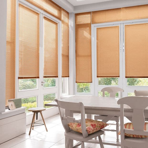 Beige pleated blinds covering tall bay windows and French doors, fully lowered and filtering warm daylight; bright dining nook with white wooden table and chairs, floral cushions, and garden view below.