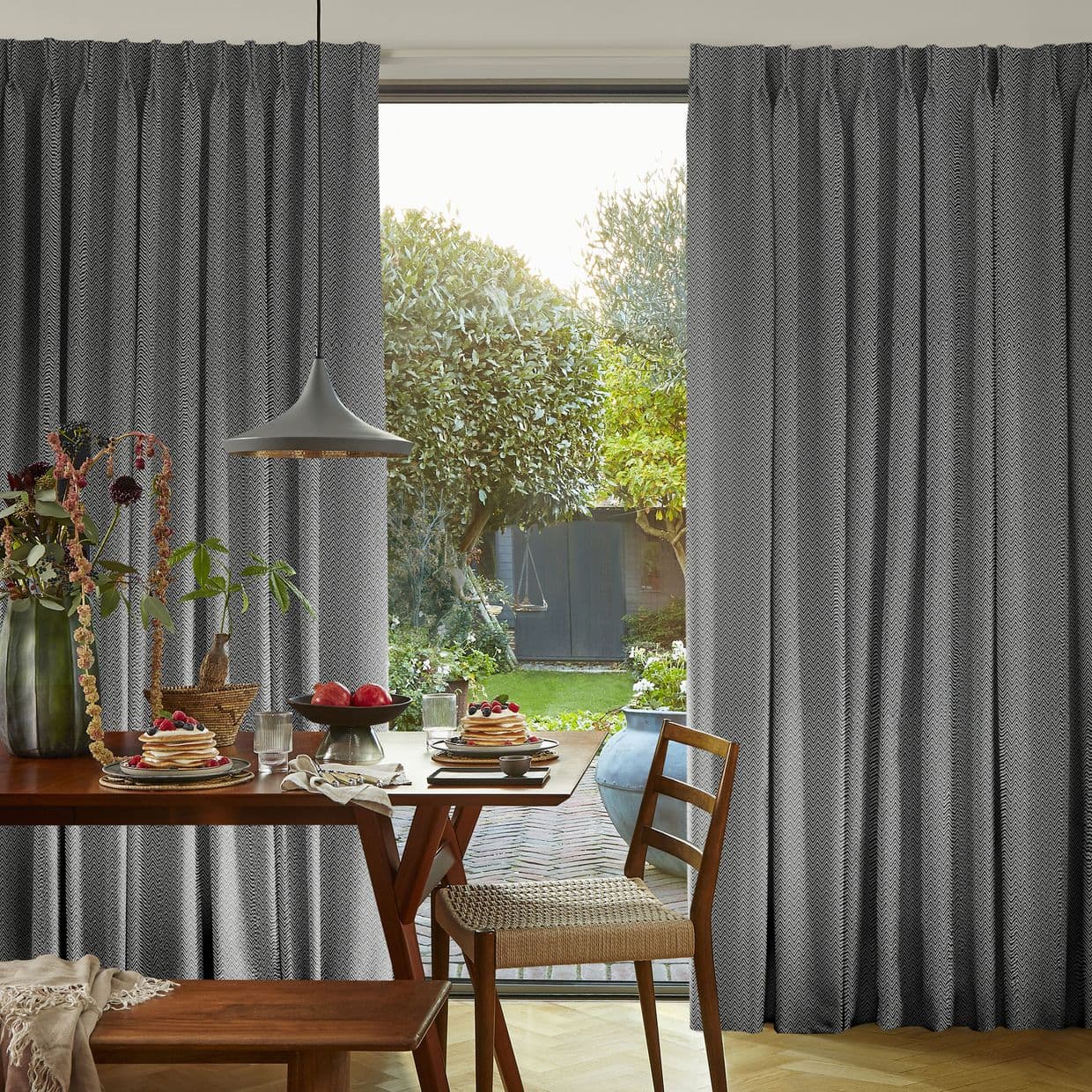 Full-length grey chevron-patterned pleated curtains pulled aside, framing a sliding glass door; they reveal morning light and a small green garden seen from a cozy dining area with wooden table.