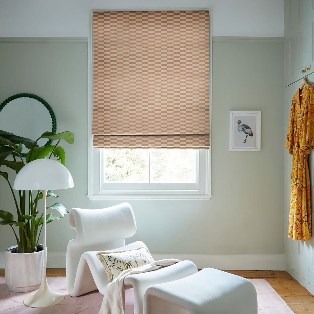 Peach-and-cream checkered Roman blind, fully lowered over the window, filtering soft daylight into a pale-green sitting nook furnished with a white sculptural lounge chair, floor lamp, plant, and hanging robe.
