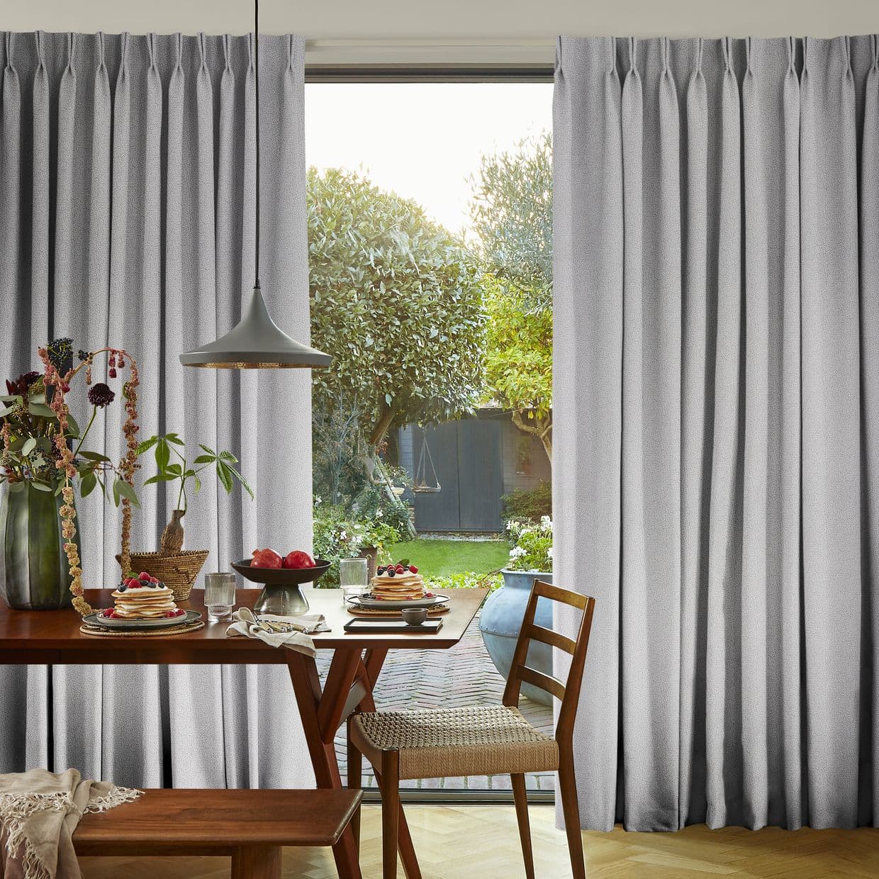 Grey, floor-length pleated curtains framing a sliding glass door, slightly parted to admit soft daylight â€” wooden dining table set for breakfast with pancakes, pendant lamp, and garden view.