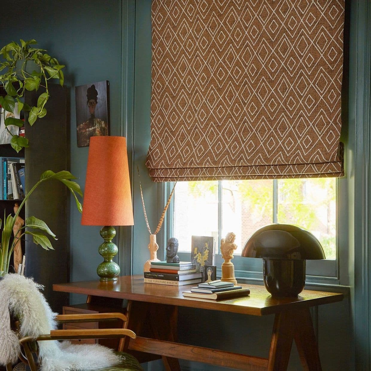 Brown Roman blind with white diamond pattern, lowered and filtering daylight over a window; set above a wooden desk with lamps, books, sculptures, teal walls and houseplants in a study.