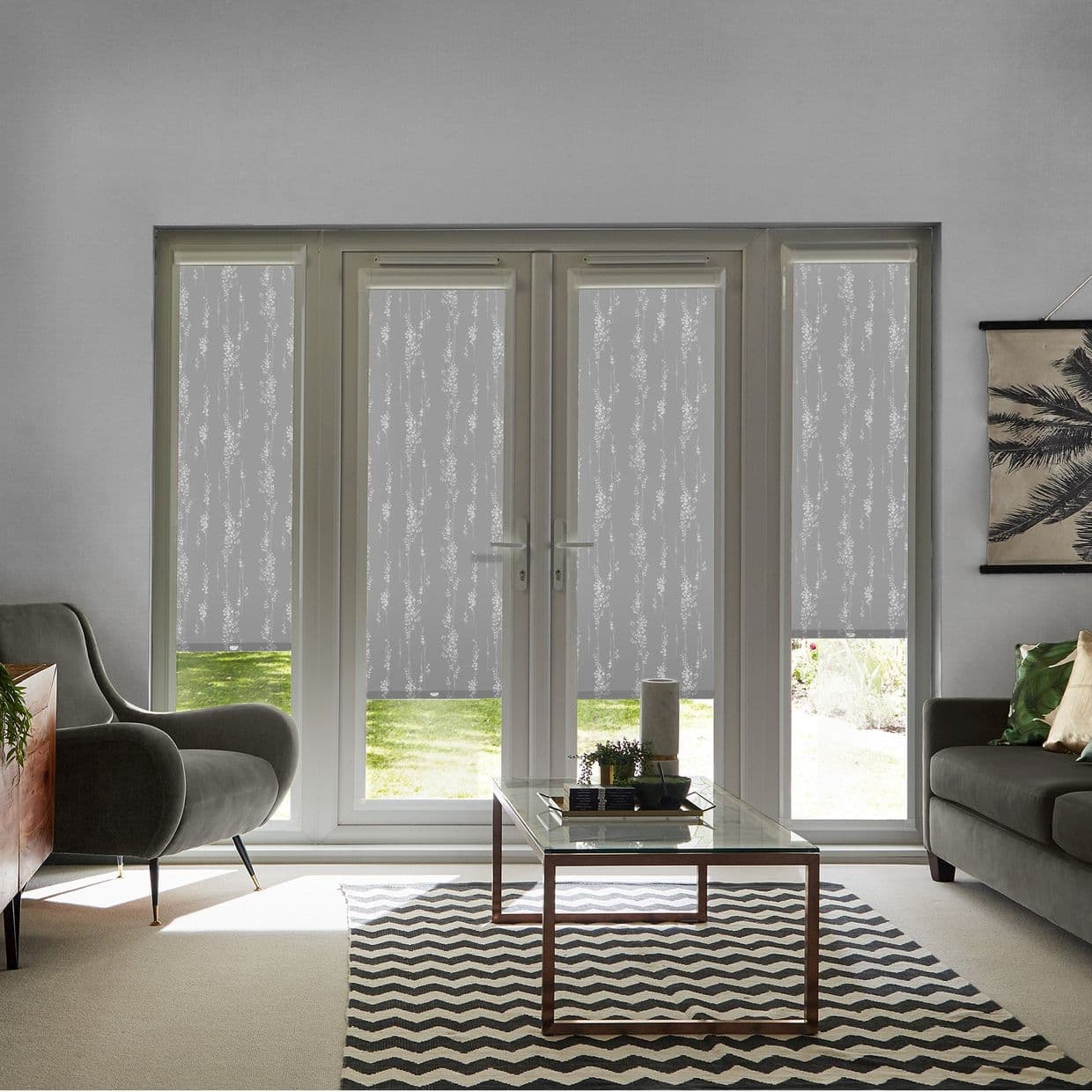 Patterned roller blinds (four panels) pulled down across French doors and sidelights, translucent vine motif filtering daylight; modern living room with grey sofas, glass coffee table and chevron rug.