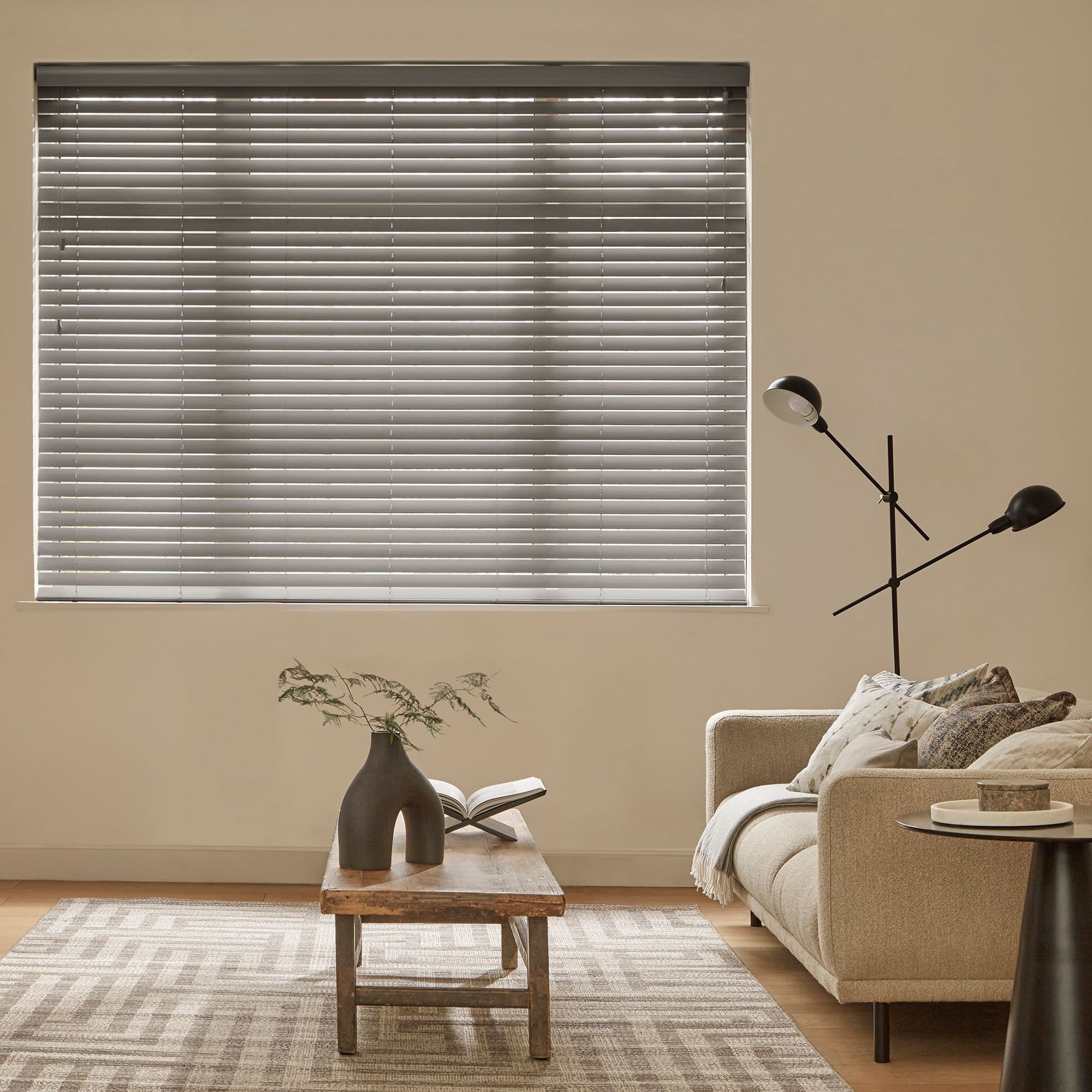 Horizontal venetian blinds covering a large wall window, slats mostly closed and filtering soft daylight; beige living room with sofa, wooden coffee table, patterned rug and black floor lamp.