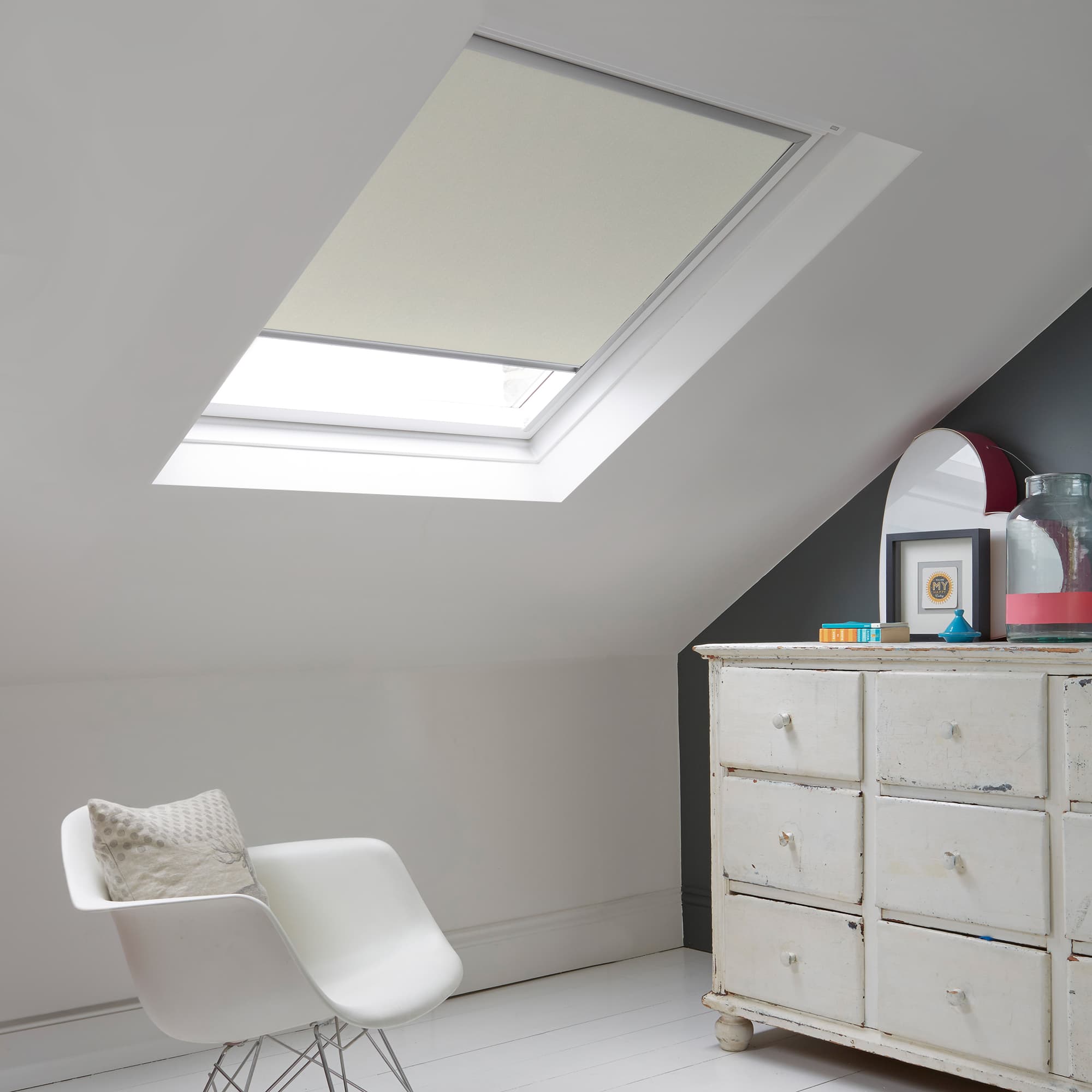 Room with white eaves wall, dark blue-grey wall to right with a shabby chic white drawer unit, a contemporary white chair, white painted floorboards and a skylight blind in off-white.