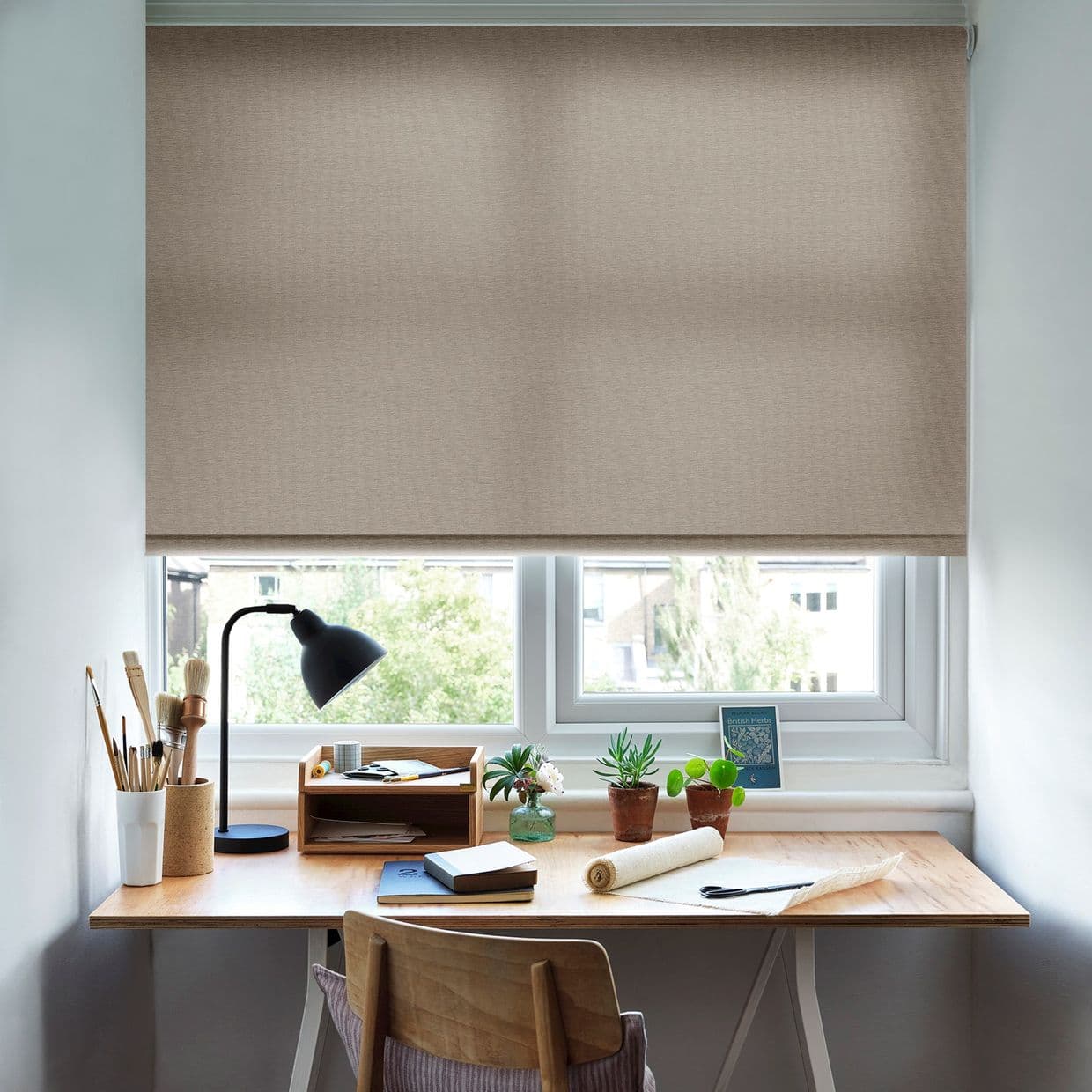 Beige fabric roller blind, fully lowered with subtle woven texture, filtering soft daylight; a wooden desk beneath holds a black lamp, paintbrushes, notebooks and small potted succulents.
