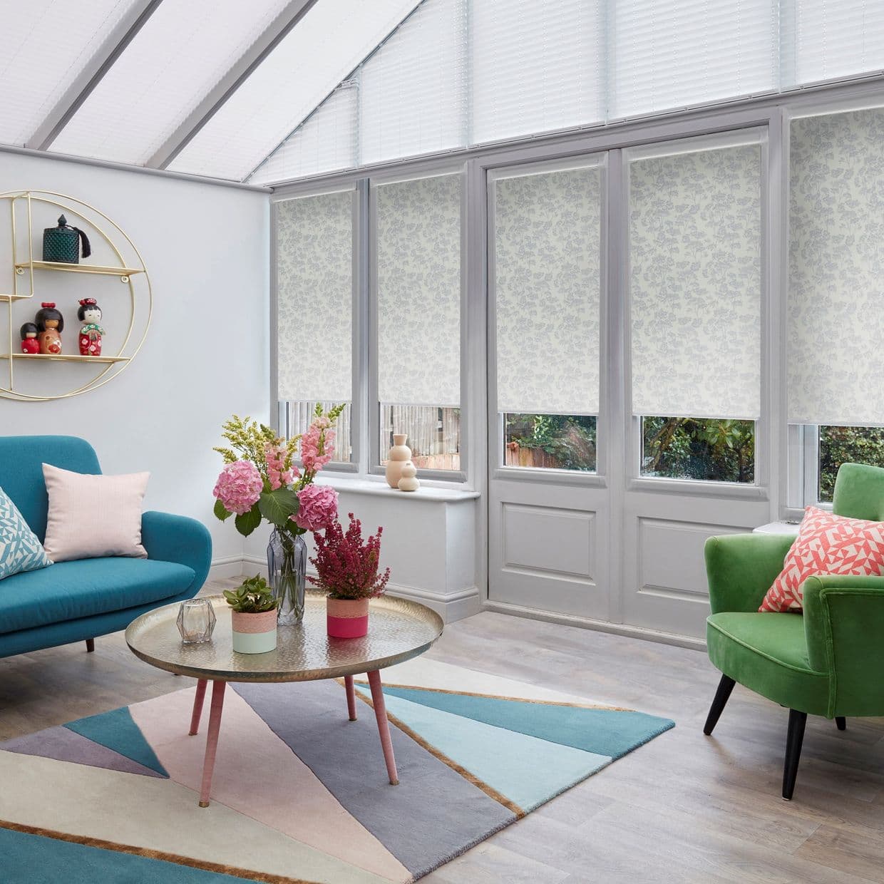 Patterned roller blinds lowered over tall panes, with pleated skylight blinds above, filtering soft daylight; conservatory-style living area with teal sofa, green armchair, round coffee table, flowers and garden view.