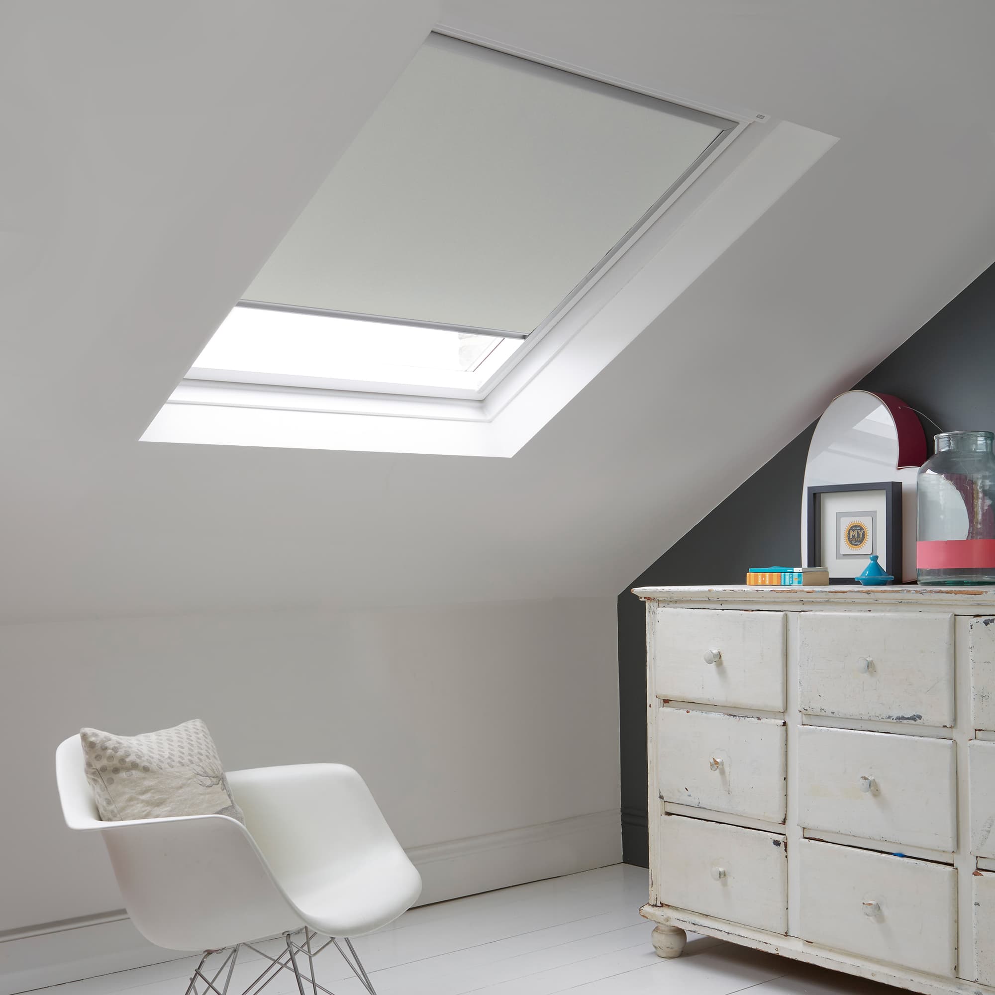 Room with white eaves wall, dark blue-grey wall to right with a shabby chic white drawer unit, a contemporary white chair, white painted floorboards and a skylight blind in pale steel grey.