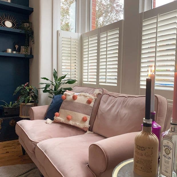 White half-height louvered plantation shutters on bay windows, closed and filtering soft daylight; cozy living room with a pink velvet sofa, pomâ€‘pom cushion, houseplants, dark shelving and candles.