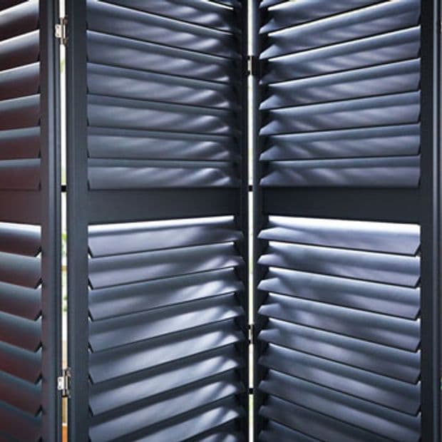 Dark three-panel louvered shutters (plantation style), hinged and partially closed, slats angled to filter thin bands of daylight into a dim interior space.