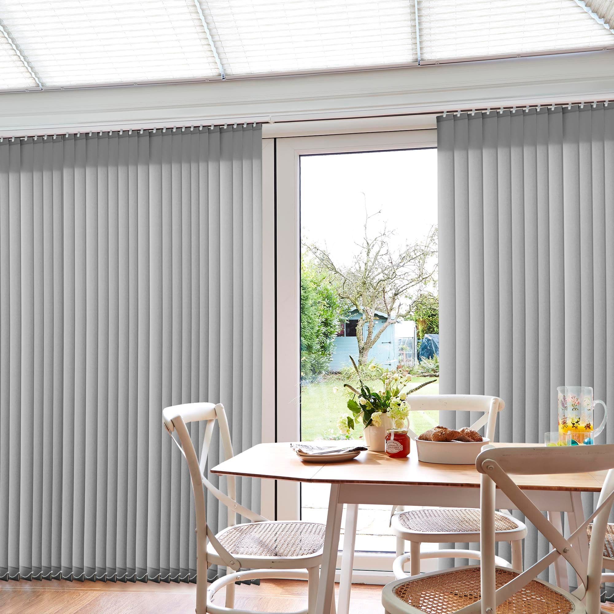 Vertical grey blinds, full-height slats on a ceiling track, drawn closed with a central gap exposing a sunlit garden; dining table with chairs, vase, pitcher and bread in foreground.