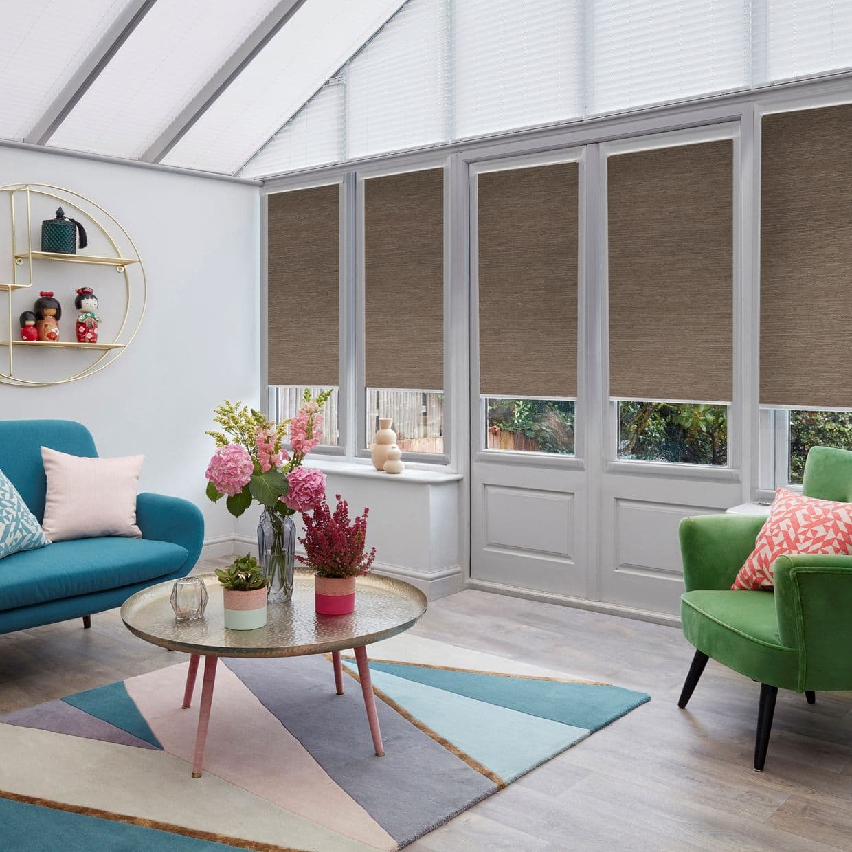 Brown fabric roller blinds, lowered to mid-height across tall conservatory windows and doors, partially filtering light; a bright sunroom with a teal sofa, green armchair, pastel rug, and floral centerpiece.