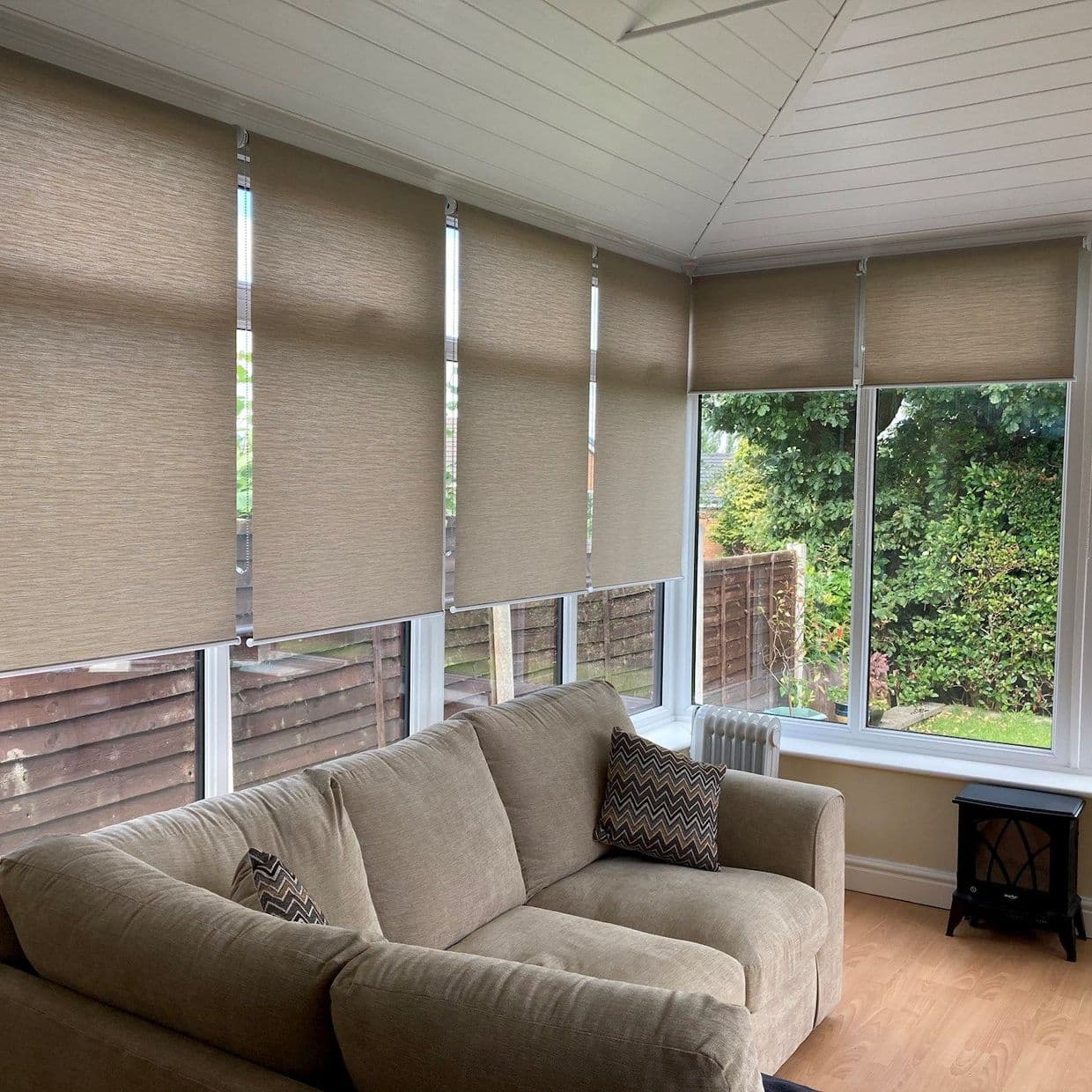Beige roller blinds covering a row of tall windows, partially lowered with slim vertical gaps; filtering daylight into a sunroom/conservatory with a beige sofa, wooden floor, and garden view.