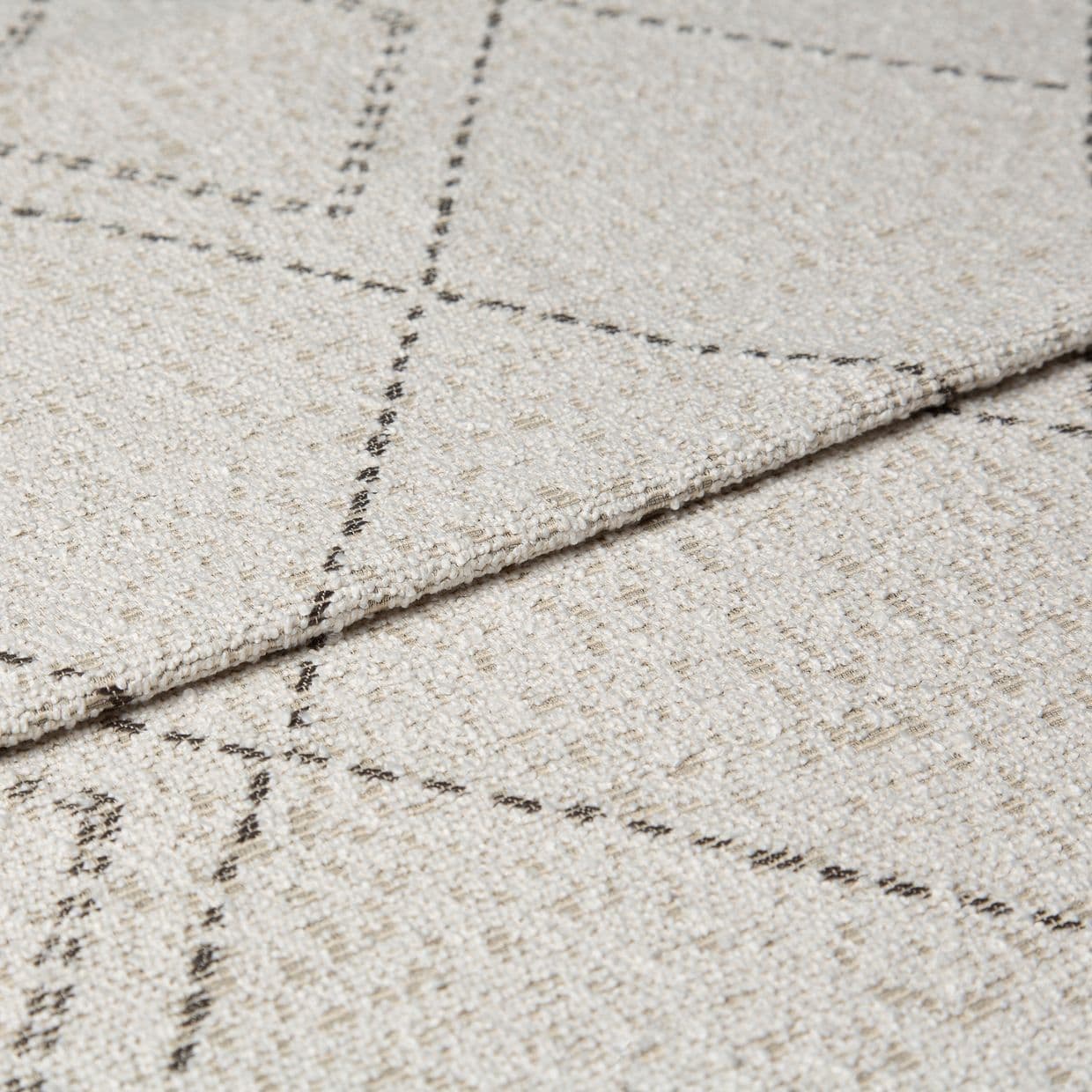 Beige woven rug with thin dashed dark grid lines, folded at the edge to show layered thickness and looped texture; close-up studio shot with neutral lighting on a flat surface.