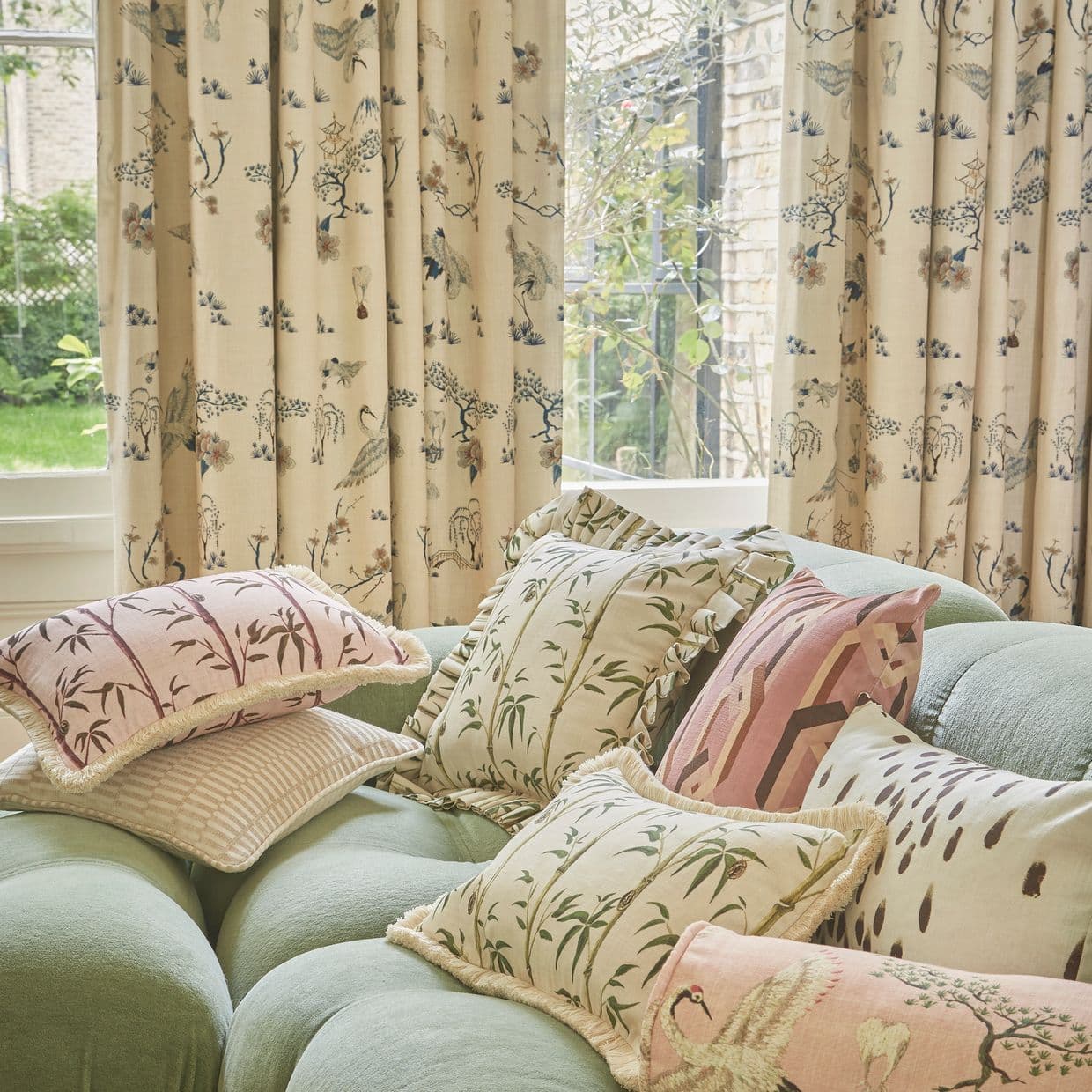 Cream pleated curtains patterned with blue-green birds and pagodas, drawn across a window softly filtering daylight; living-room corner with tufted pale-green sofa piled with decorative cushions, garden beyond.