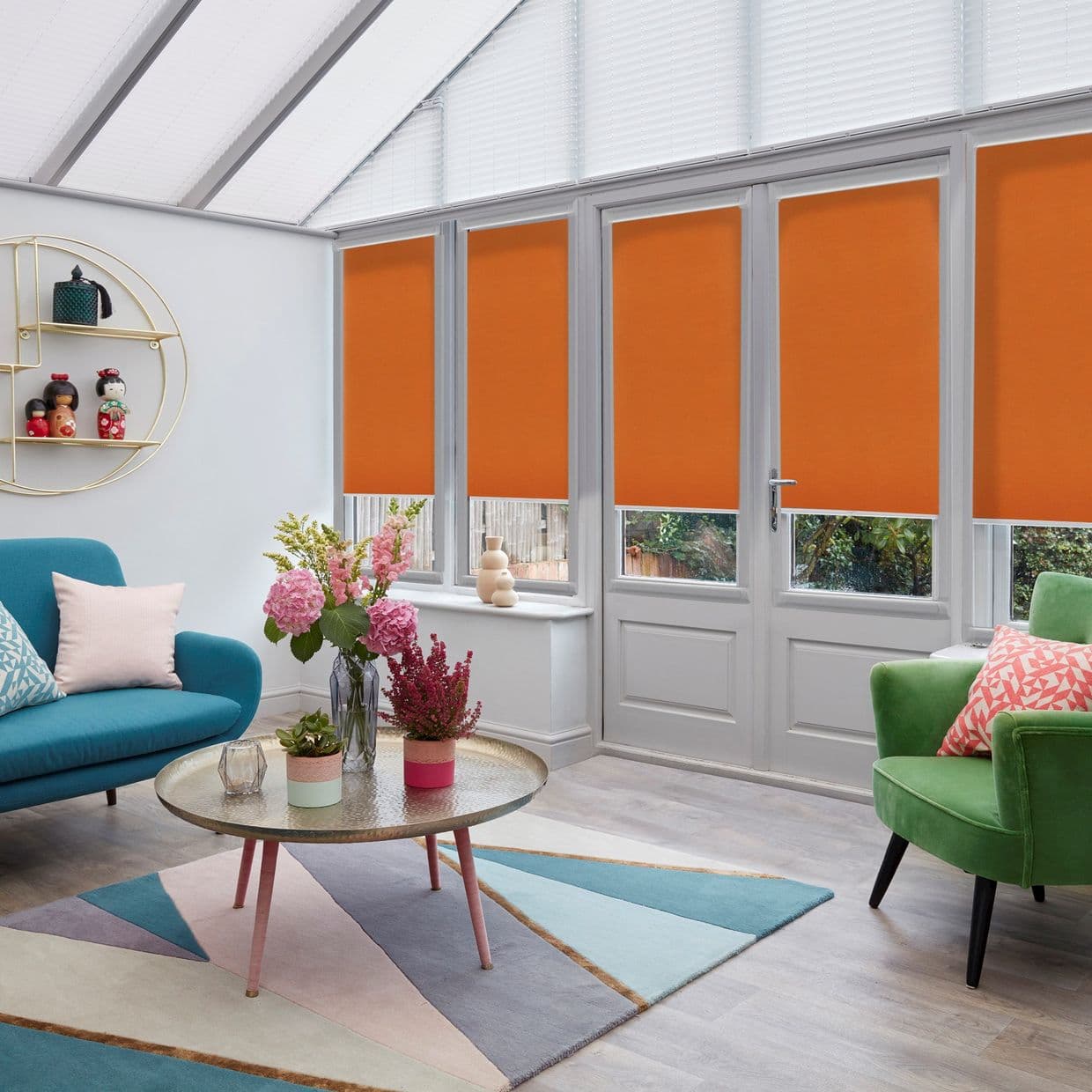 Orange roller blinds on tall glazed doors are partially lowered, covering upper glass while leaving short lower panes exposed, in a bright conservatory-style living room with colorful sofas, coffee table.