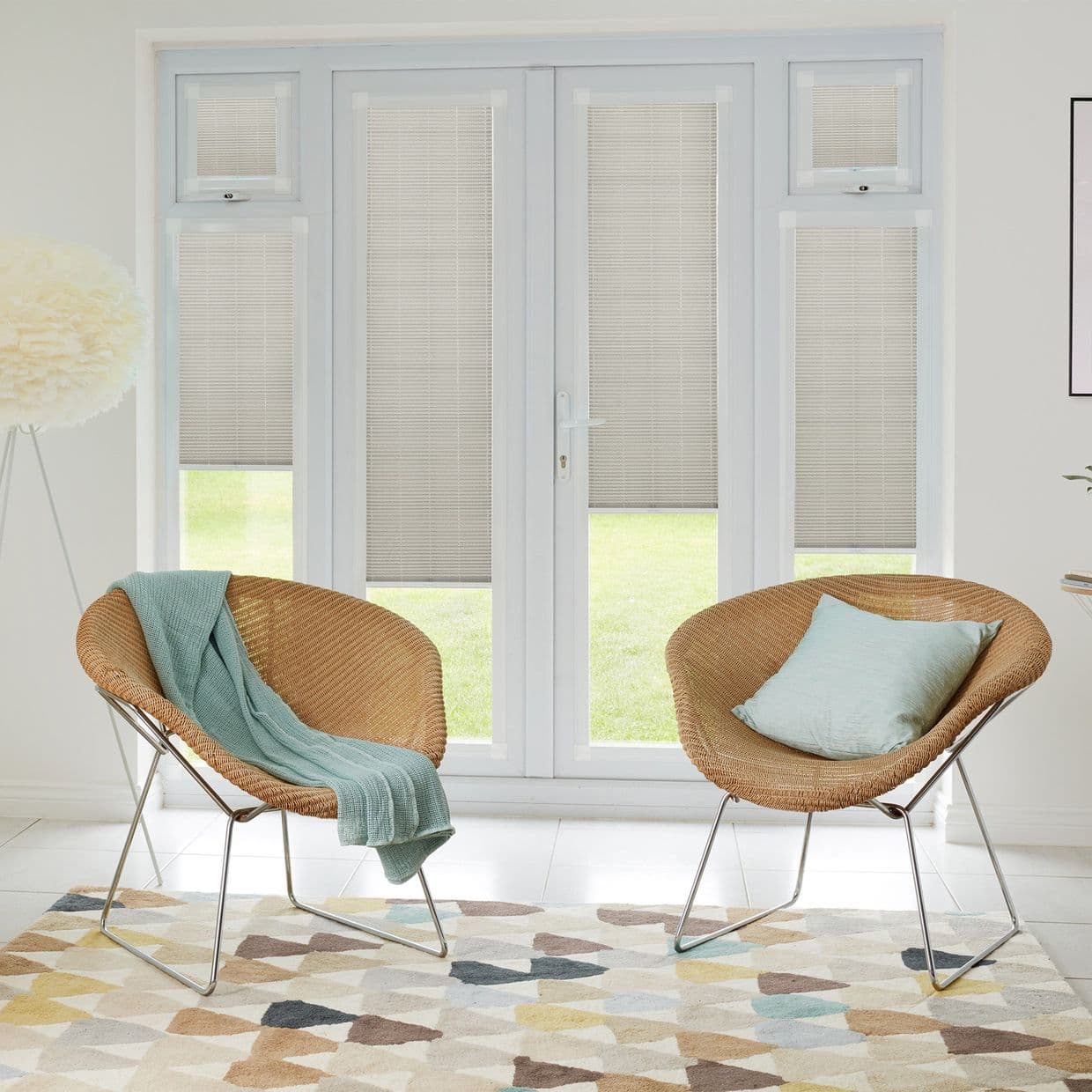 Beige pleated blinds fitted to white-framed French doors and sidelights, partially lowered and filtering daylight; modern bright sitting area with two wicker chairs, teal throw, patterned rug, lawn outside.