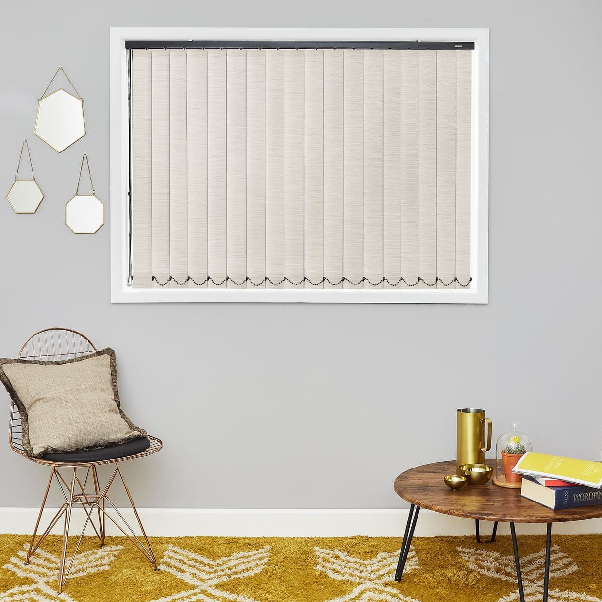 Beige vertical blinds, closed with beaded chain at hem, filtering light; in a gray-walled living room over a mustard rug, beside a wire chair, hanging mirrors, and a small wooden table.