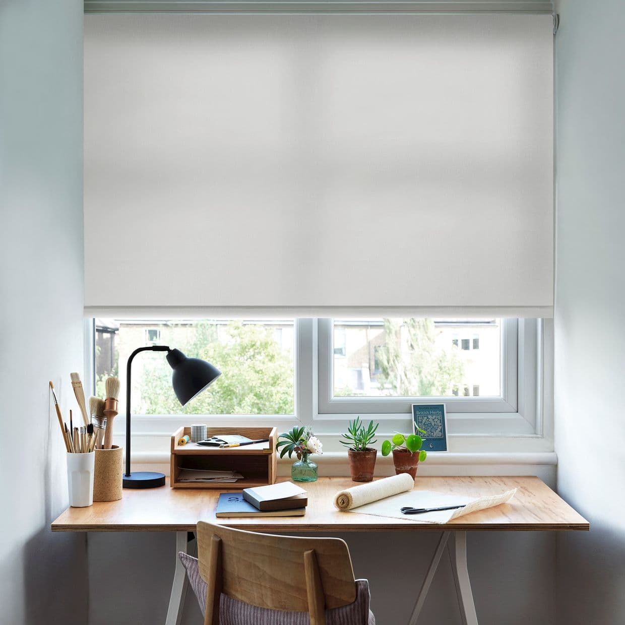 White fabric roller blind, lowered to just above sill, softly diffusing daylight; wooden desk beneath with lamp, brushes, notebooks and small potted plants, bright home office with windowed suburban view.