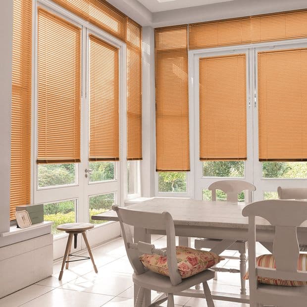 Orange horizontal Venetian blinds cover tall bay windows and glass doors, lowered to filter warm sunlight into a bright dining nook with white wooden table and chairs, overlooking a garden.