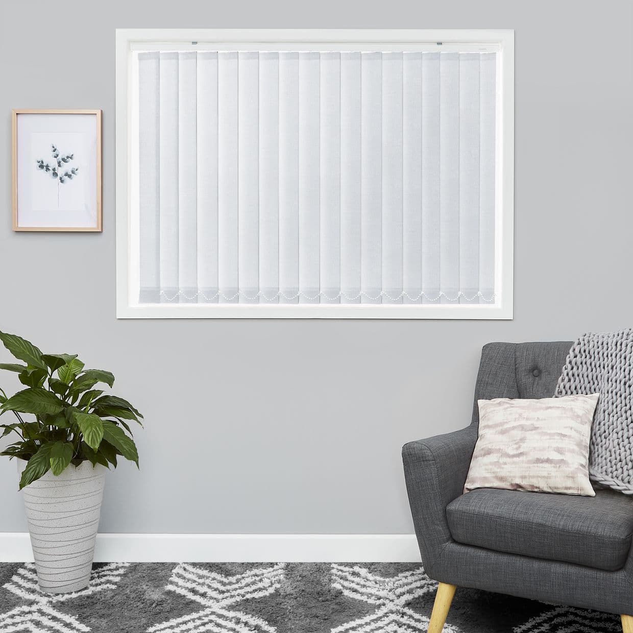 White vertical blinds covering a window, narrow fabric slats with scalloped bottoms; closed and diffusing soft daylight; living-room setting with grey armchair, knitted throw, potted plant, framed art.