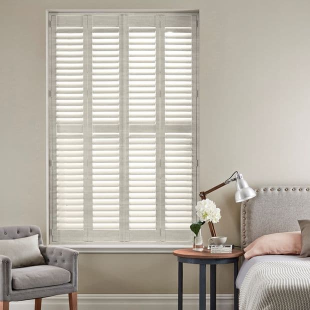 White plantation shuttersâ€”four tall louvered panelsâ€”fully closed, softly filtering daylight into a neutral bedroom corner with upholstered headboard, bedside table, lamp, vase of flowers and a gray armchair.