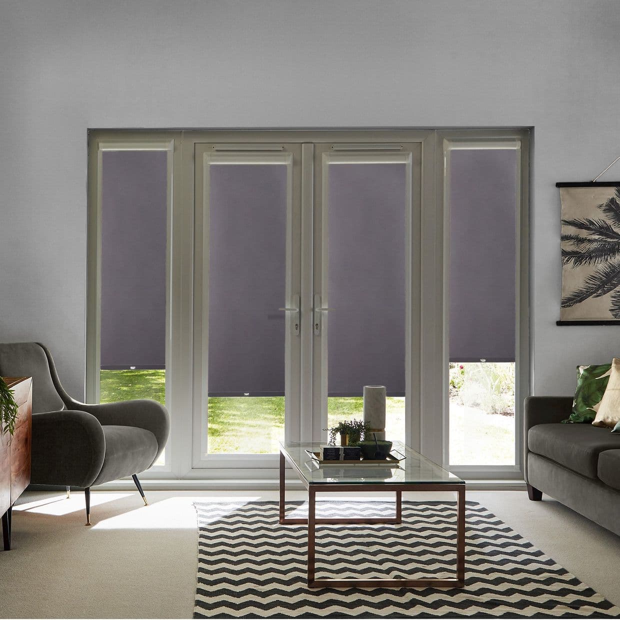 Grey roller blinds on French doors and sidelights, lowered mid-height and blocking most light; reveal narrow garden strip; modern living room with glass coffee table, zigzag rug, armchair and sofa.