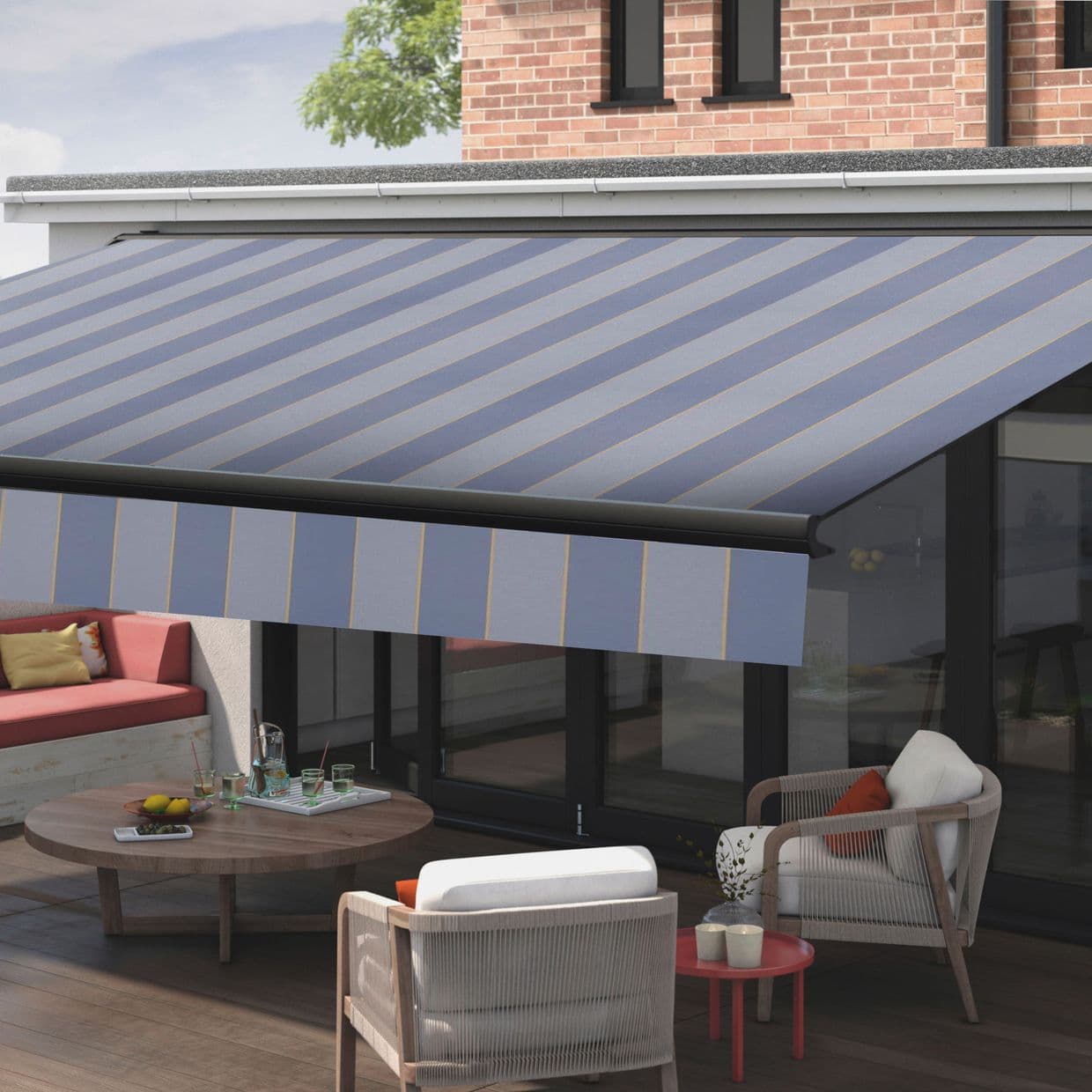 Striped retractable fabric awning with blue-gray diagonal stripes, fully extended over a patio; shading glass doors and outdoor seating â€” daytime, brick upper facade, wooden deck, lounge chairs and coffee table.