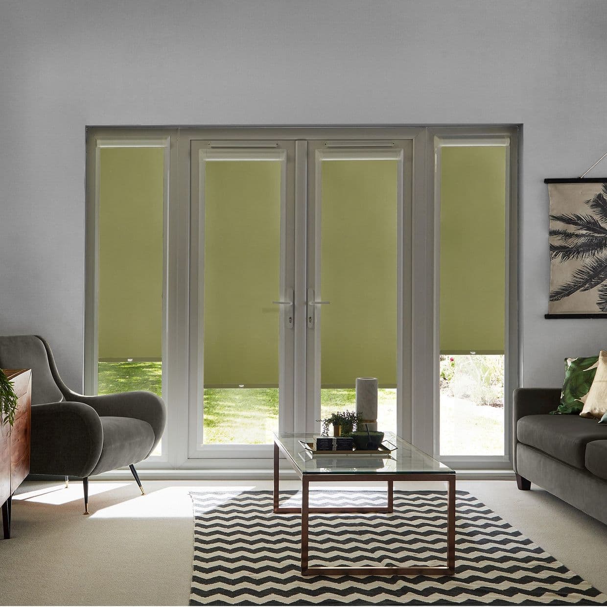 Olive roller blinds lowered across double French doors and sidelights, blocking upper light while revealing lawn at bottoms; modern living room with grey sofas, glass coffee table, zigzag rug, daylight.