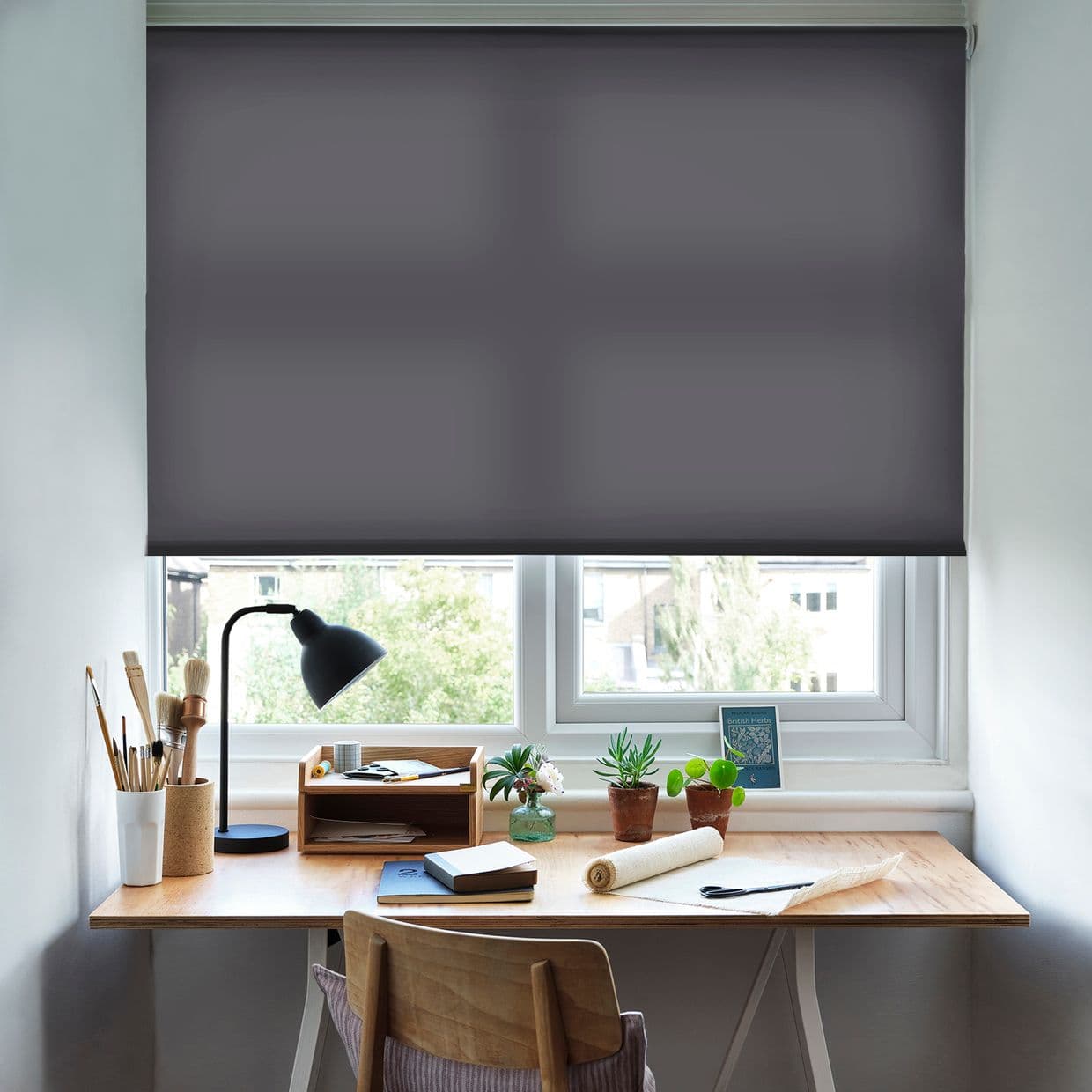 Dark grey roller blind inside the window frame, lowered almost fully and dimming daylight; above a neat wooden desk with lamp, brushes, notebooks, potted succulents, chair and treed urban view.