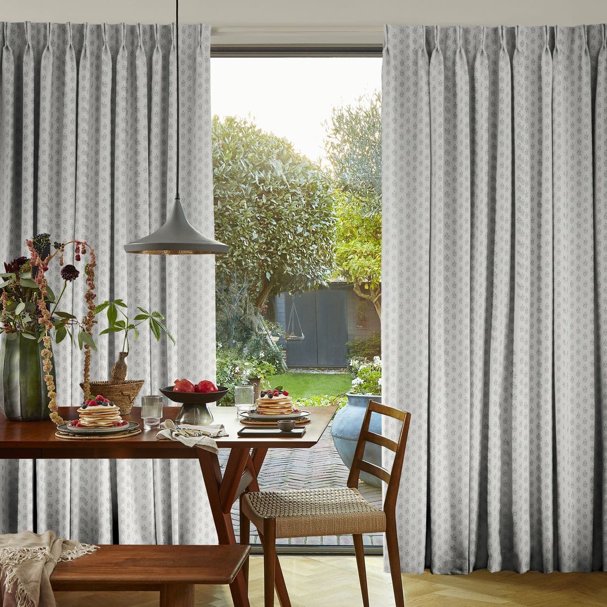 Patterned pleated curtains, pale grey, pulled aside revealing a sliding glass door; filtering soft daylight into a dining nook with wooden table set for breakfast and a green garden view.