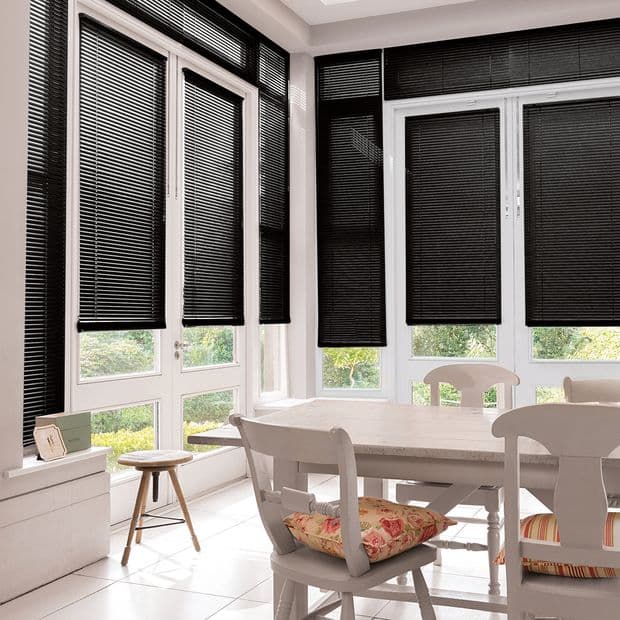 Black horizontal-slatted blinds fitted across tall white-framed doors and windows, mostly lowered and closed, filtering light; bright dining corner with white table and chairs and garden glimpses.