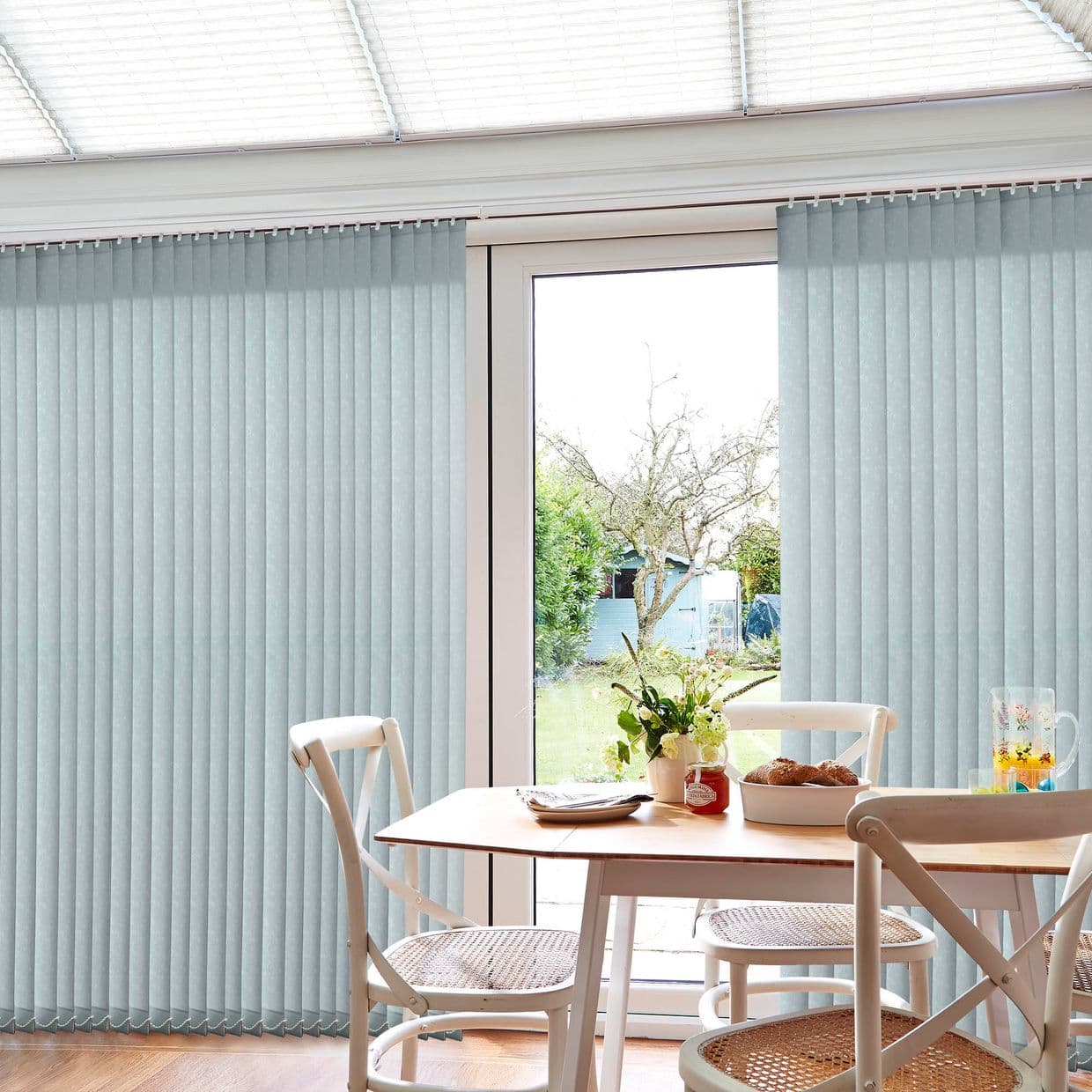 Pale blue vertical blinds, floor-to-ceiling and split to reveal a central glass door, filtering daylight; small dining table and chairs nearby, bright conservatory opening to a green garden.