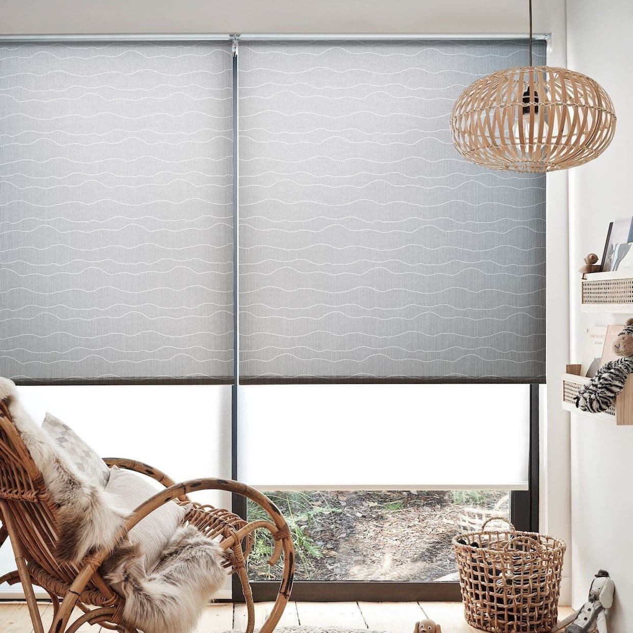 Light-gray patterned roller blinds (two adjacent panels with subtle white wavy lines), fully lowered and diffusing daylight over a sliding window; cozy nook with rattan chair, wicker pendant, basket, floor.
