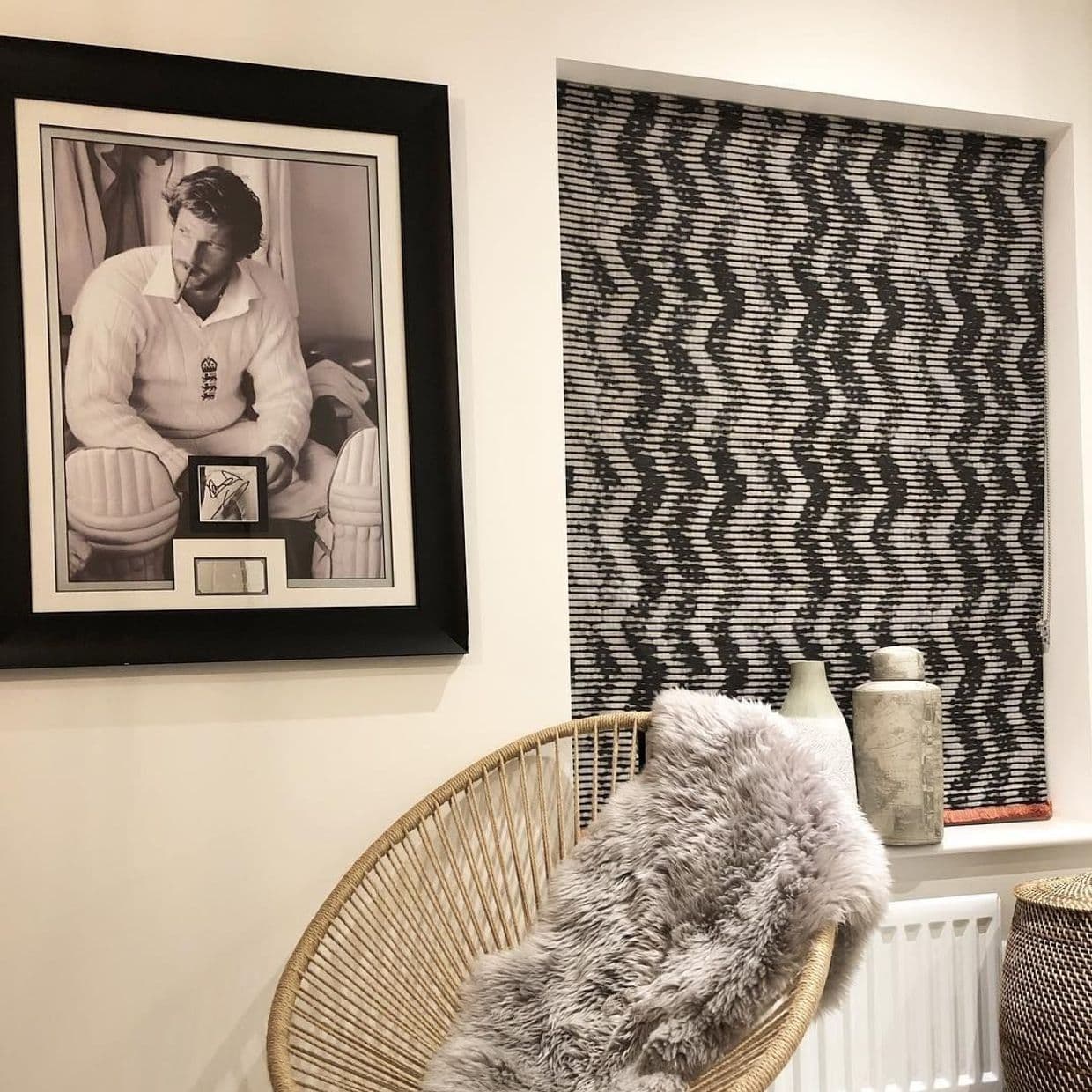 A patterned roller blind, recessed and lowered, displaying black-and-white zigzag stripes covering the window. Context: cozy corner with framed black-and-white portrait, wicker chair draped in gray fur, ceramic vases.
