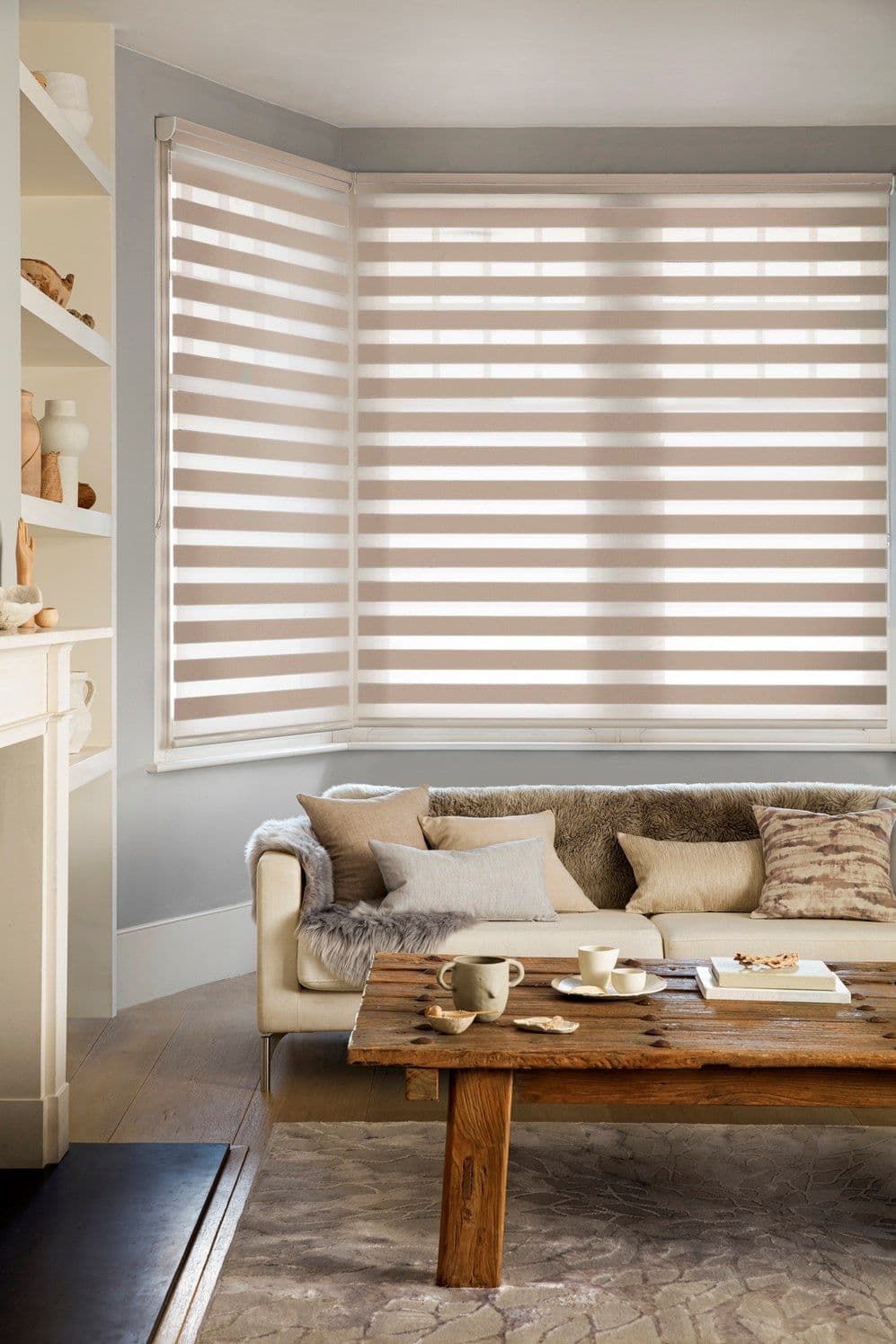 Zebra roller blinds (beige-and-white horizontal stripes) covering a three-panel bay window, lowered and filtering soft daylight; in a neutral-toned living room with sofa and rustic wooden coffee table.