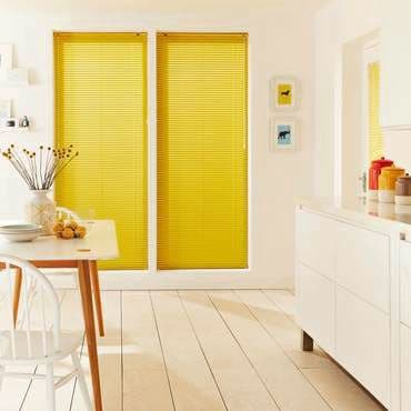 Two tall yellow pleated blinds covering side-by-side windows, fully lowered and filtering sunlight; modern bright kitchen-dining area with white furniture, wooden floor, table, vase and fruit.