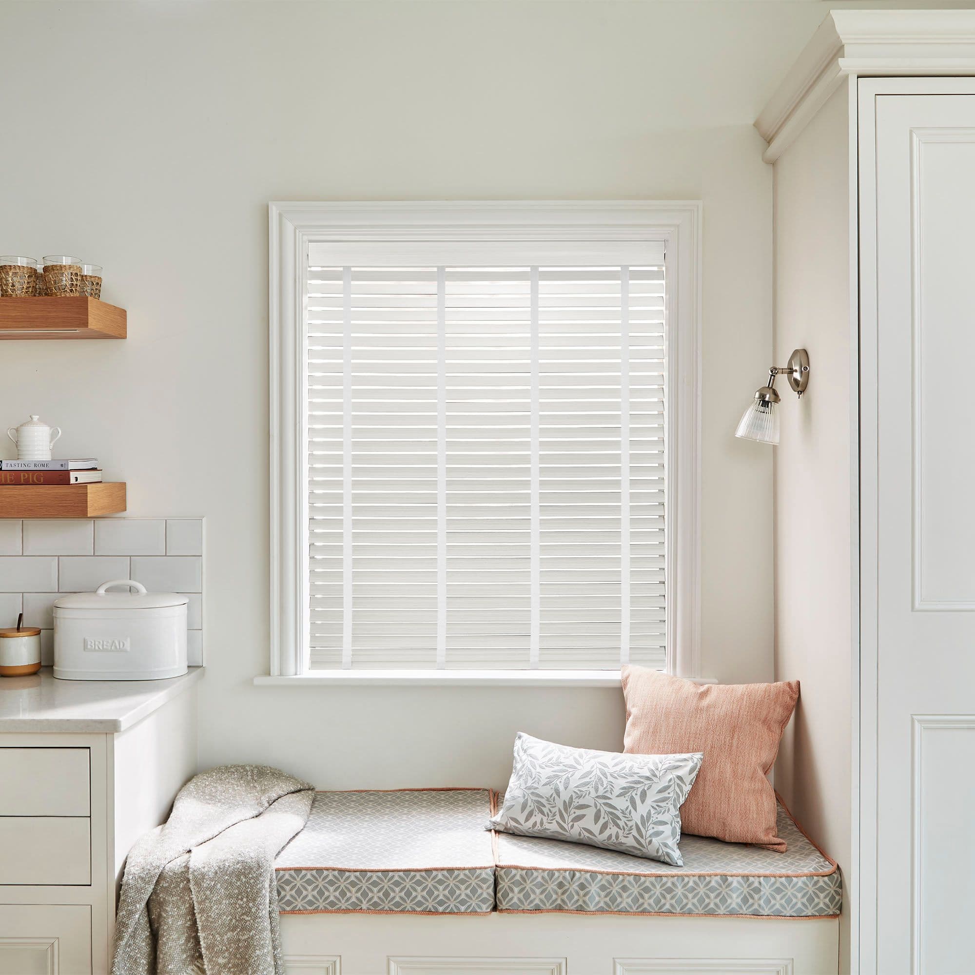 White horizontal slatted blinds in the window frame, closed and filtering soft daylight; kitchen bench nook with cushioned seat, two pillows, floating wooden shelf, countertop and bread tin labeled BREAD.
