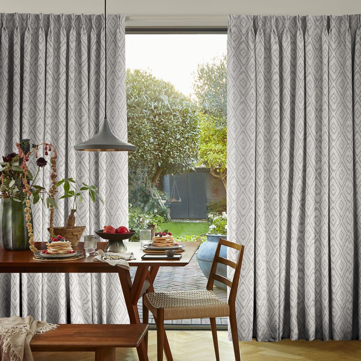 Patterned gray pleated curtains frame a sliding glass door, partly drawn to reveal a sunlit backyard; dining table with plates, pendant lamp, and wooden chairs in a cozy breakfast nook.