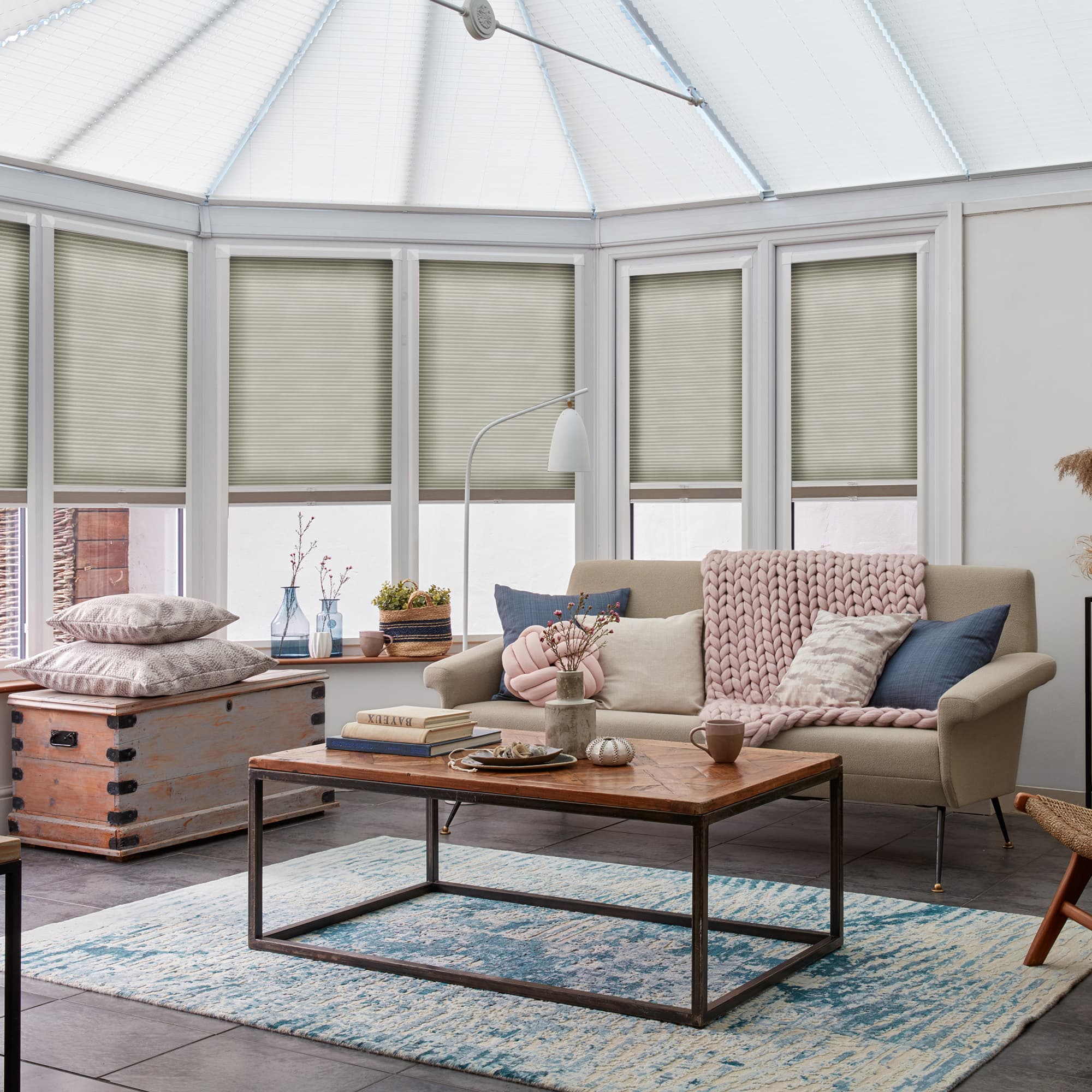 Pale beige cellular pleated blinds across bay windows, lowered to mid-height and filtering daylight; conservatory living area with sofa draped in chunky knit, wood-and-metal coffee table, decorative cushions and rug.