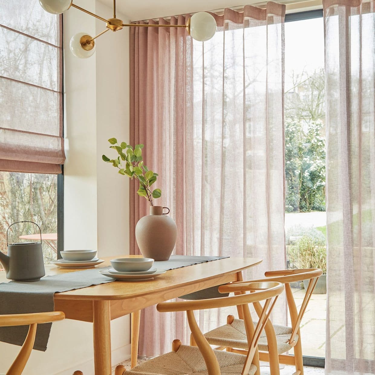 Sheer dusty-pink floor-to-ceiling curtains and matching Roman blind at left drawn, softly filtering daylight across a sliding glass door; wooden dining table with vase and woven chairs in bright corner.
