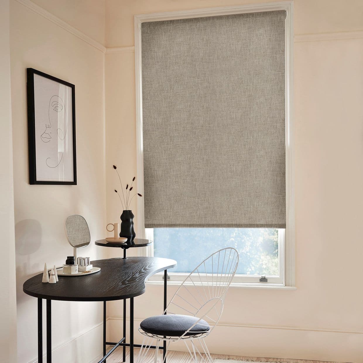 Gray textured roller blind, lowered and filtering daylight through a narrow bottom gap; hangs over a window in a pale, minimalist room with a black desk, wire chair, and framed line-drawing wall print.