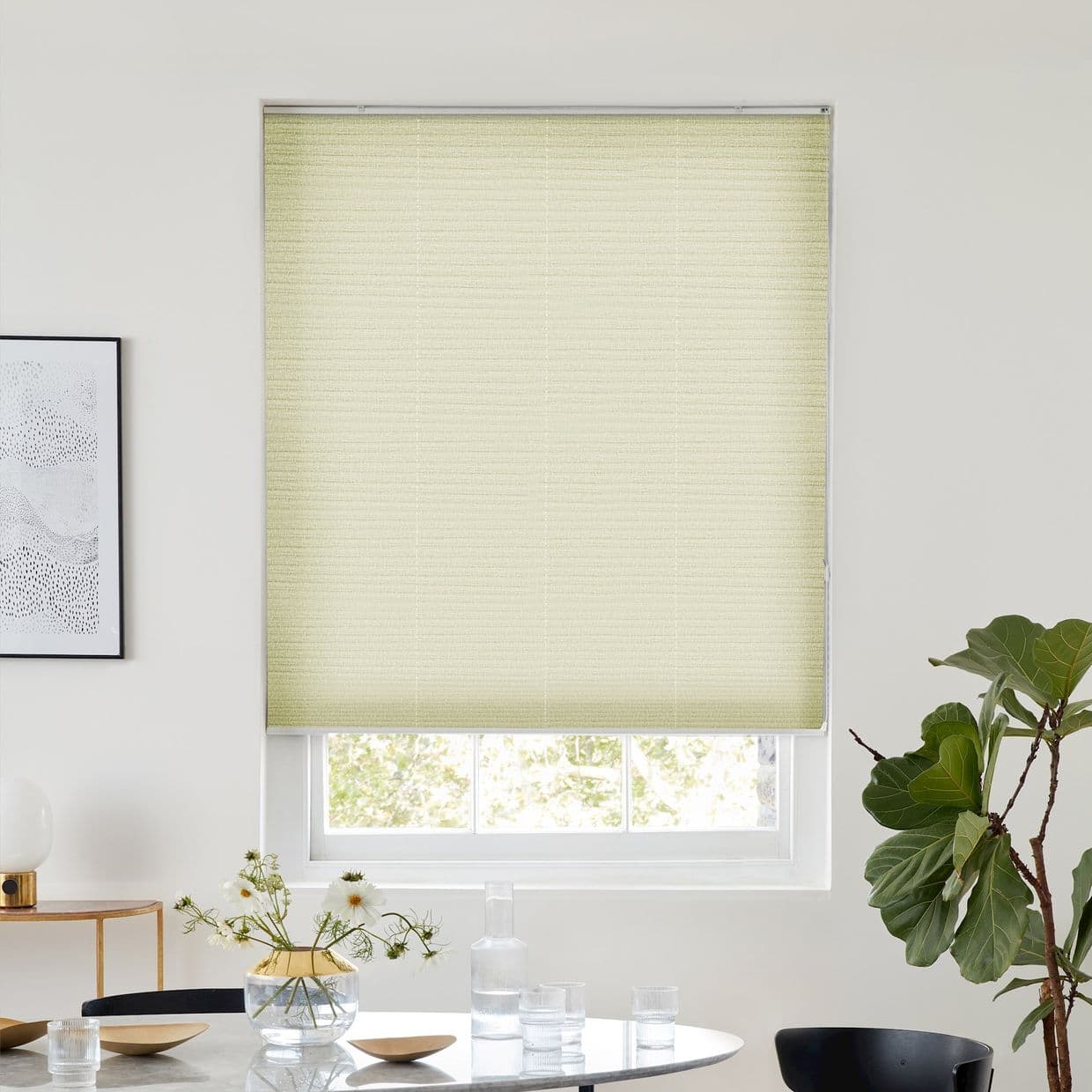 Light-yellow pleated cellular shade, fully lowered over a sash window, filtering daylight; set in a bright minimalist dining area with round table and vase, framed art left, fiddleâ€‘leaf fig right.