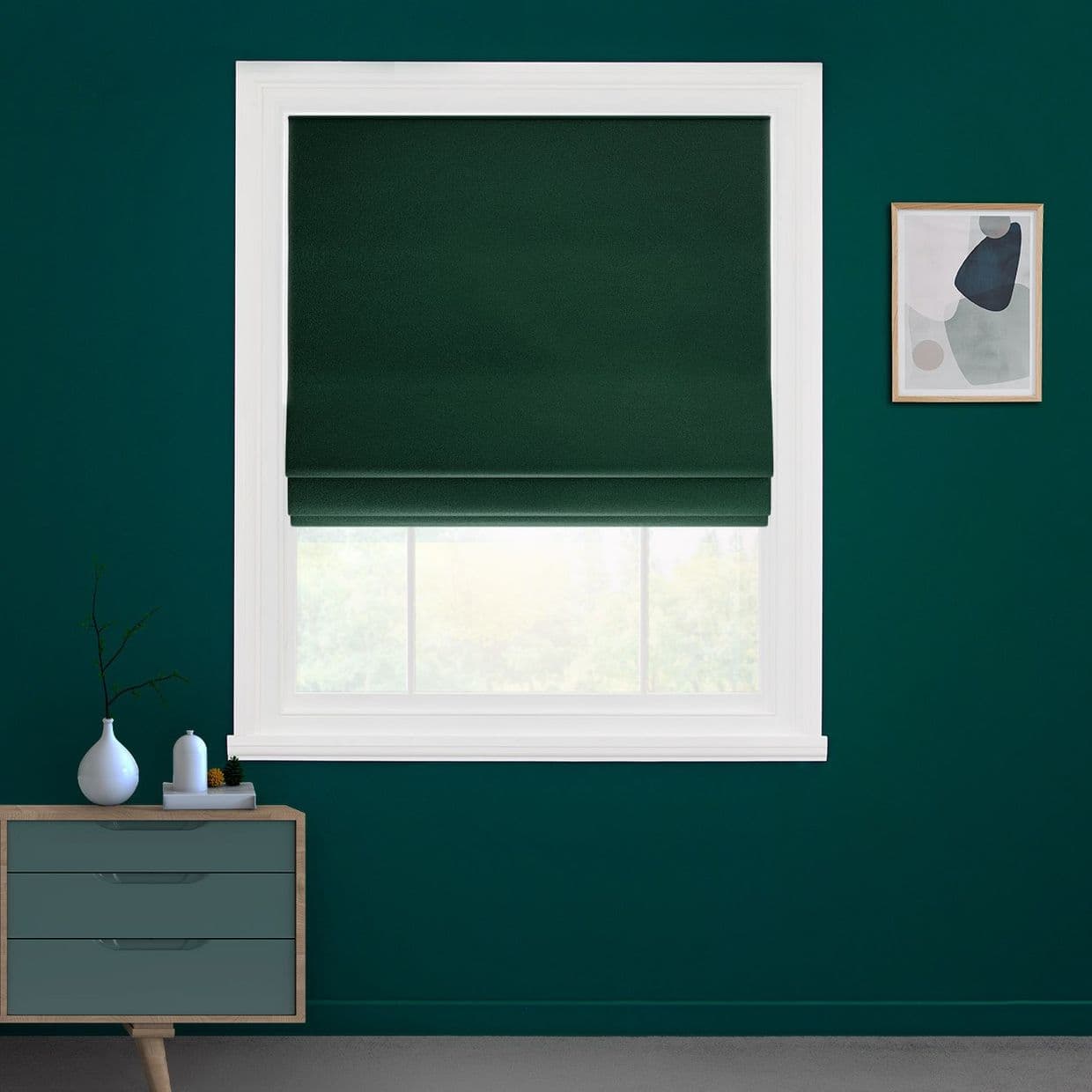 Dark green Roman blind, centered in white window frame, folded slightly near the bottom and covering most glass; teal-painted room with a small dresser left and framed abstract art right.