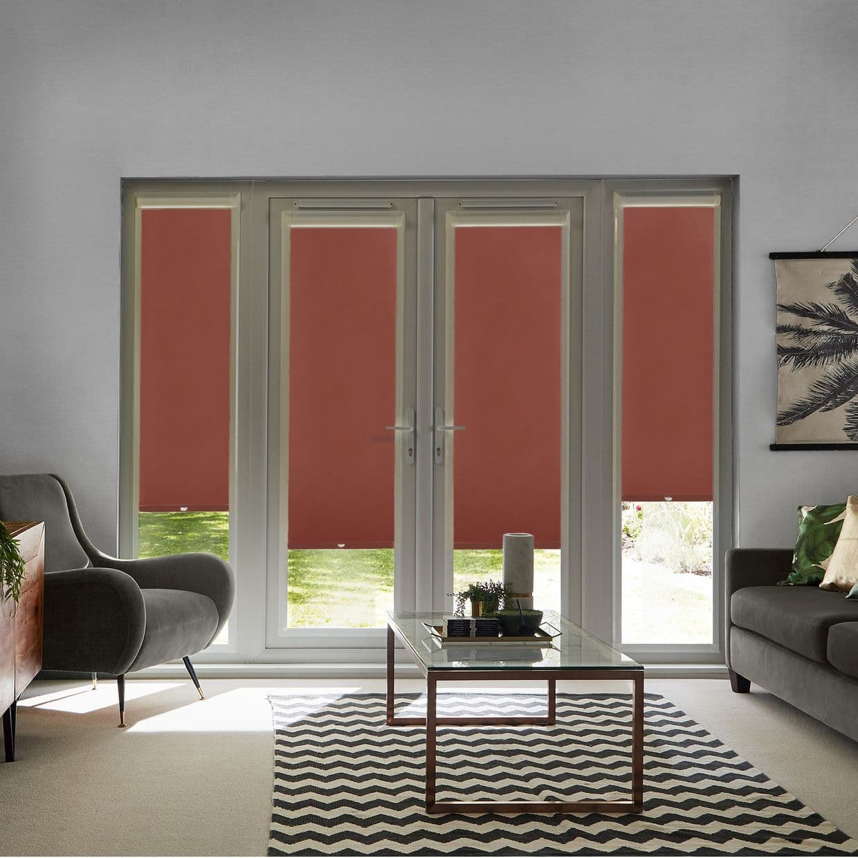 Deep-red roller blinds cover four tall French-door panels, partially lowered with narrow bottom gaps, blocking most light; modern living room with grey sofas, glass coffee table, chevron rug, lawn outside.