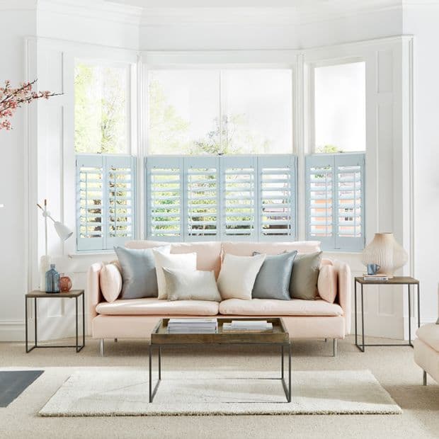Pale-blue cafÃ©-style plantation shutters cover the lower halves of a bay window; slatted panels partially open, filtering daylight into a bright living room with pastel sofa, cushions and coffee table.