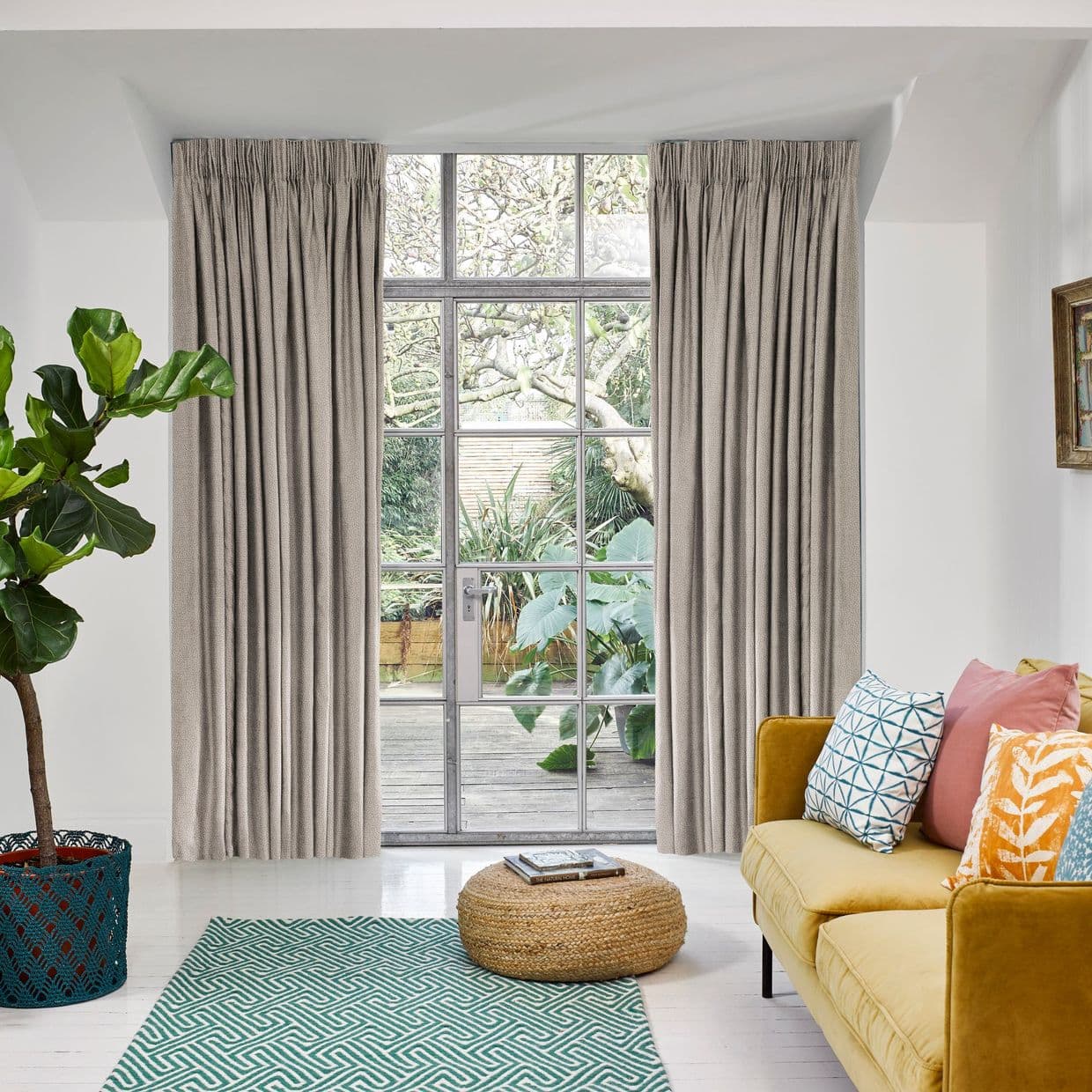 Floor-to-ceiling pleated beige curtains, pulled slightly aside to frame a central metal-framed glass door, letting daylight and a leafy garden view into a bright living room with mustard sofa.