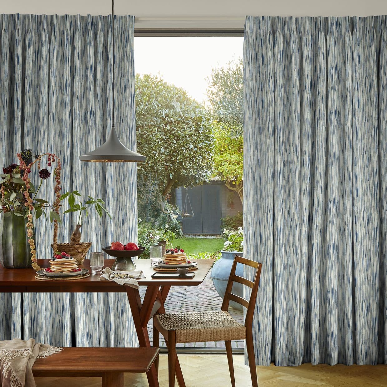 Floor-to-ceiling curtains with blue-grey vertical brushstroke pattern, parted to reveal a sunlit garden and sliding glass door. Inside, a wooden dining table set for breakfast beneath a pendant lamp.