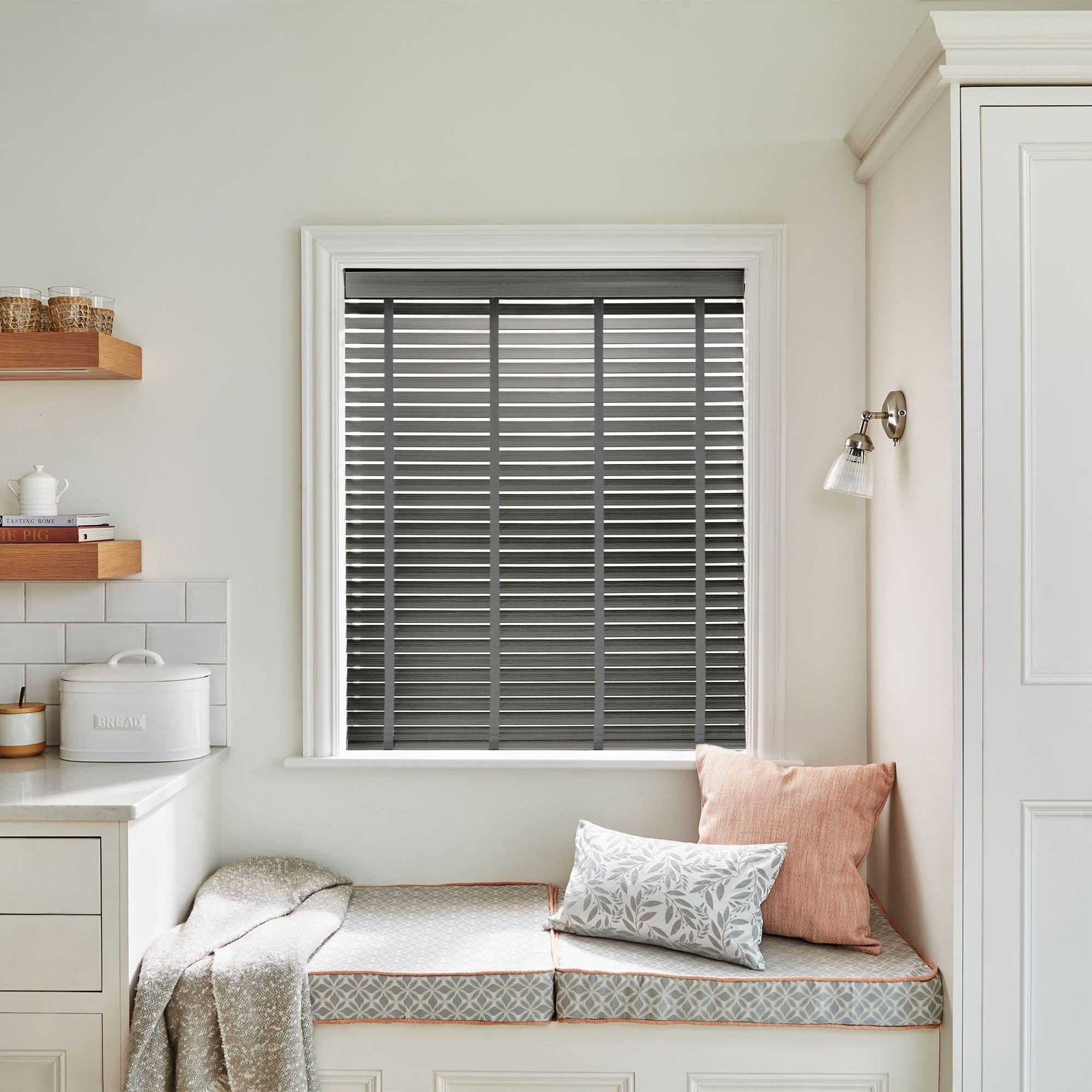 Gray horizontal Venetian blinds fully covering the window, slatted and closed with narrow light gaps; they filter daylight above a cushioned window seat in a neutral, softly lit kitchen nook.
